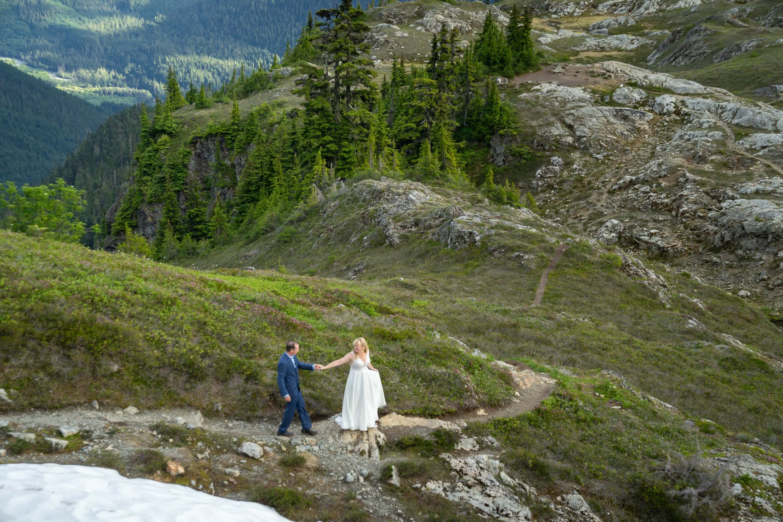 A bride and groom walking through a lush subalpine meadow in the North Cascades with purple heather and blooming wildflowers. The bride is wearing a wedding dress and the groom is a suit, and there are rocks and snow in the meadow around them.