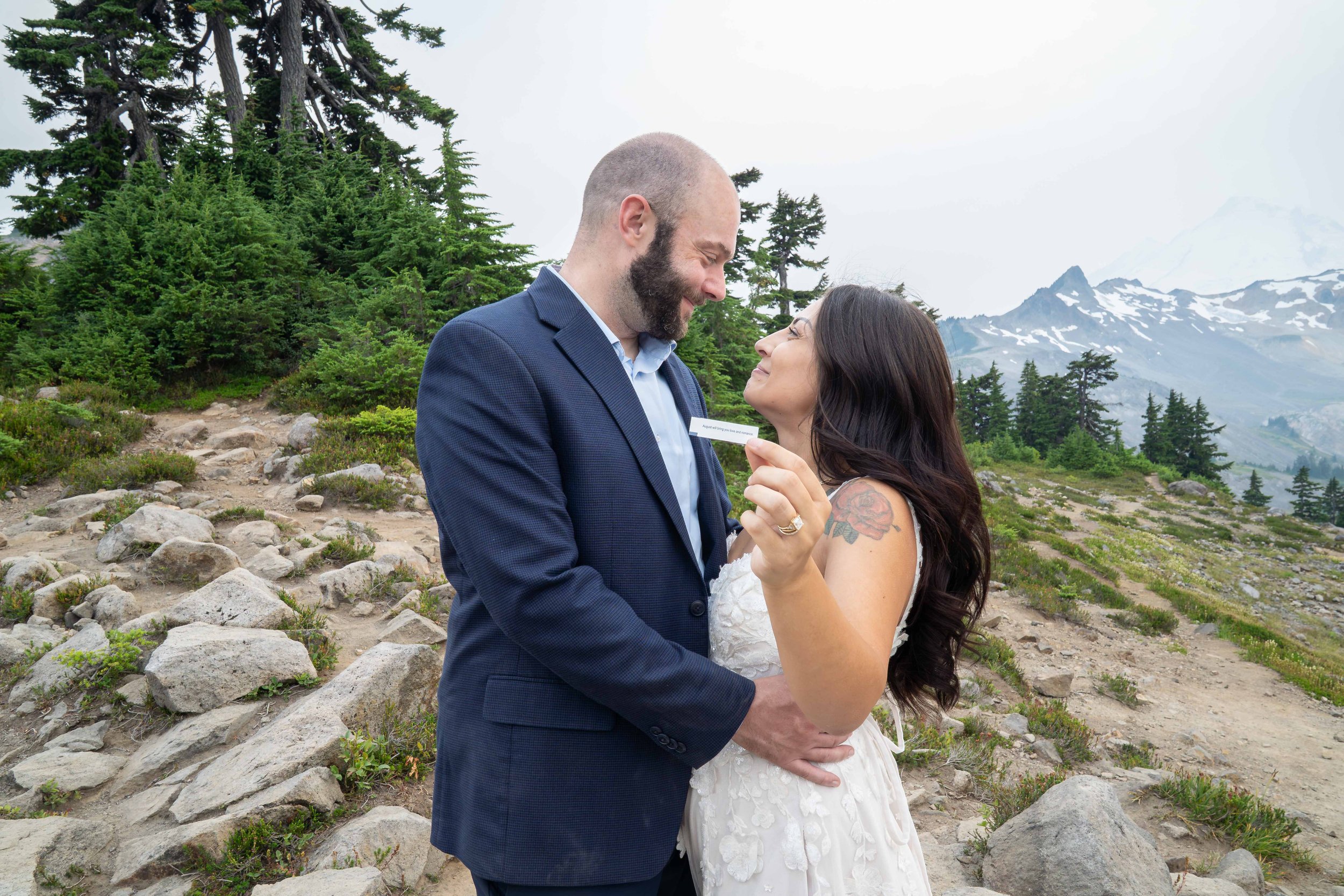 A bride and groom standing together during their Mt. Baker elopement on Ptarmigan Ridge while the bride holds up a fortune that says "August will bring you love". Coleman Pinnacle and Mt. Baker are visible in the background.