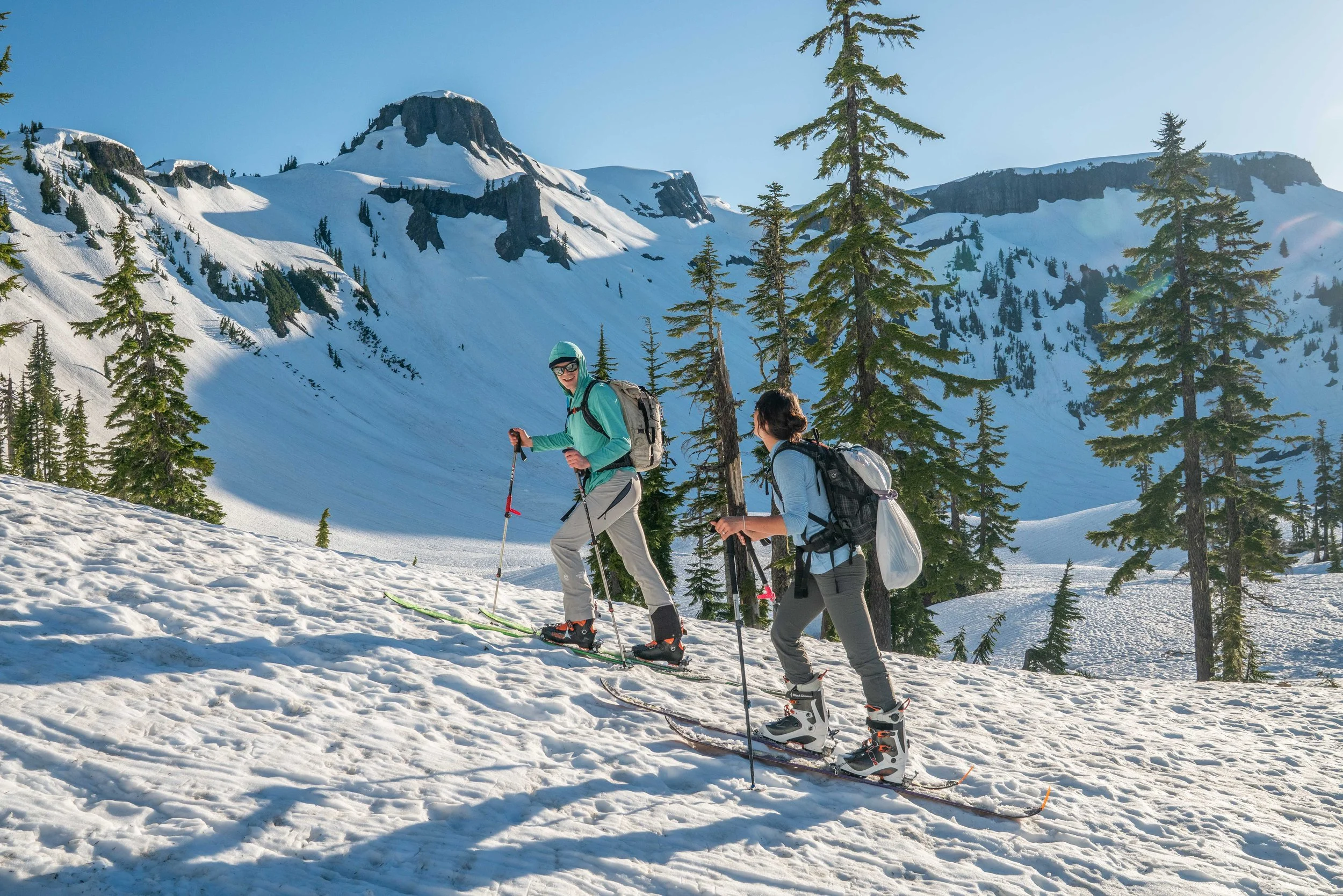 A couple skinning uphill on skis in the Baker Backcountry. The woman has a wedding dress strapped to her backpack.