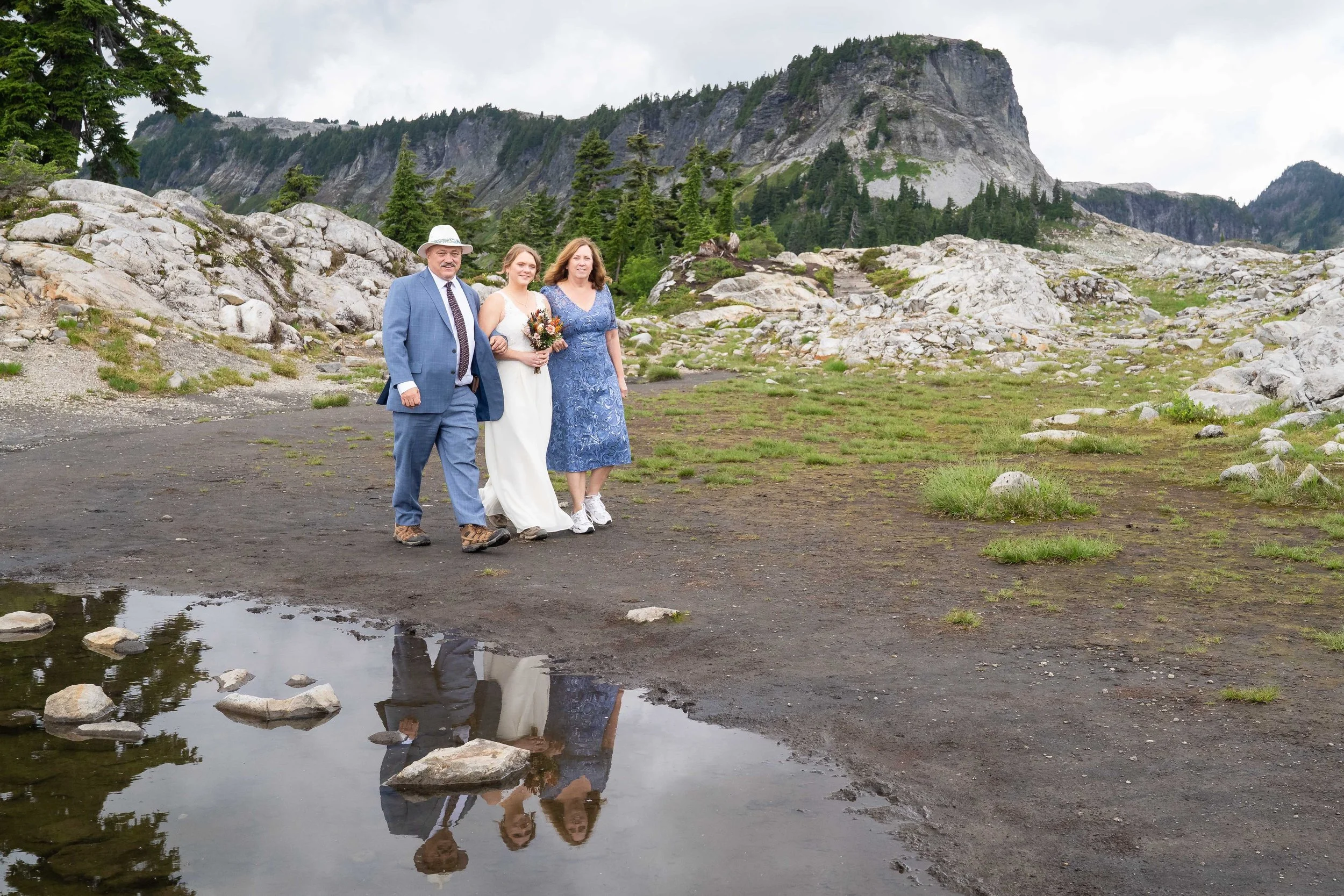Photo of a bride and her parents walking across an alpine meadow at Artist Point as part of a wedding ceremony, with a small lake in the foreground and Table Mountain in the background.