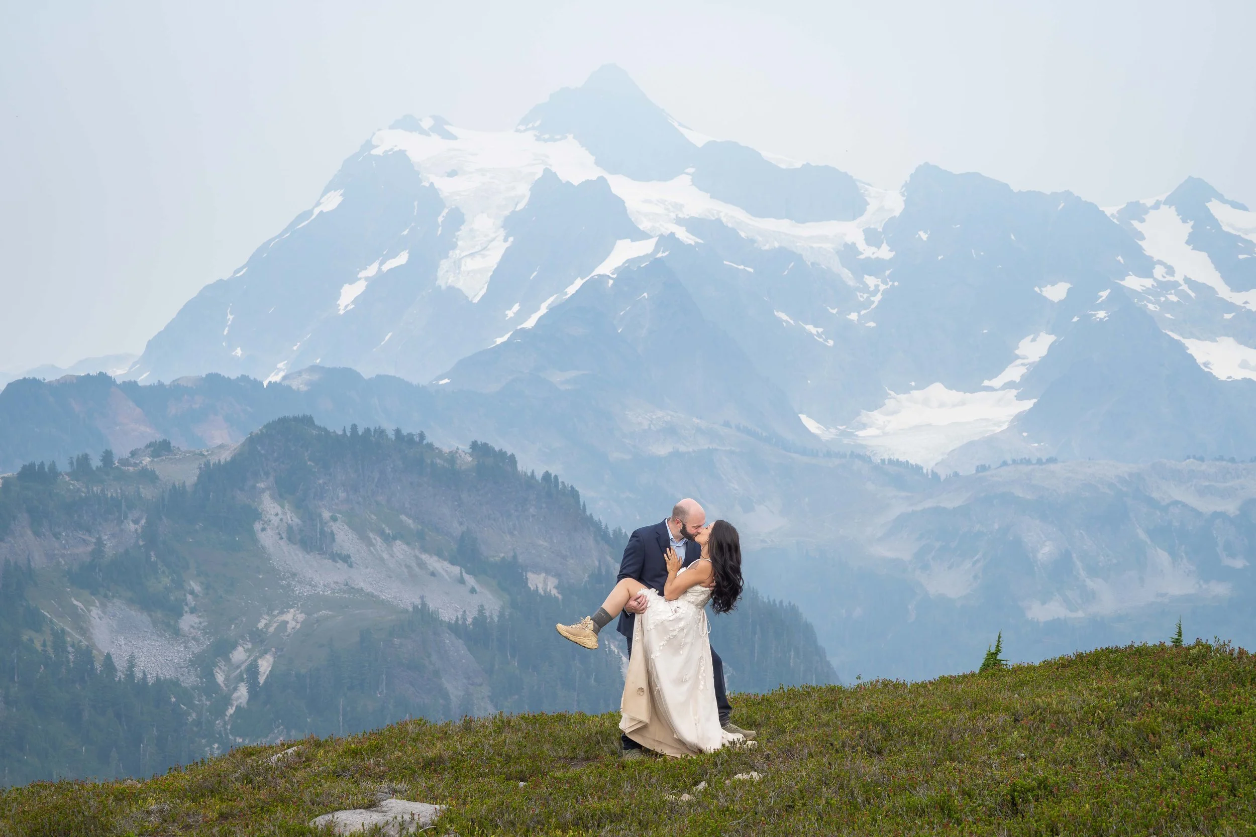 A groom holds his bride and tilts her back for a kiss during a Mt. Baker adventure wedding at Ptarmigan Ridge. They are standing in a blooming heather meadow and Mt. Shuksan is visible directly behind the couple.