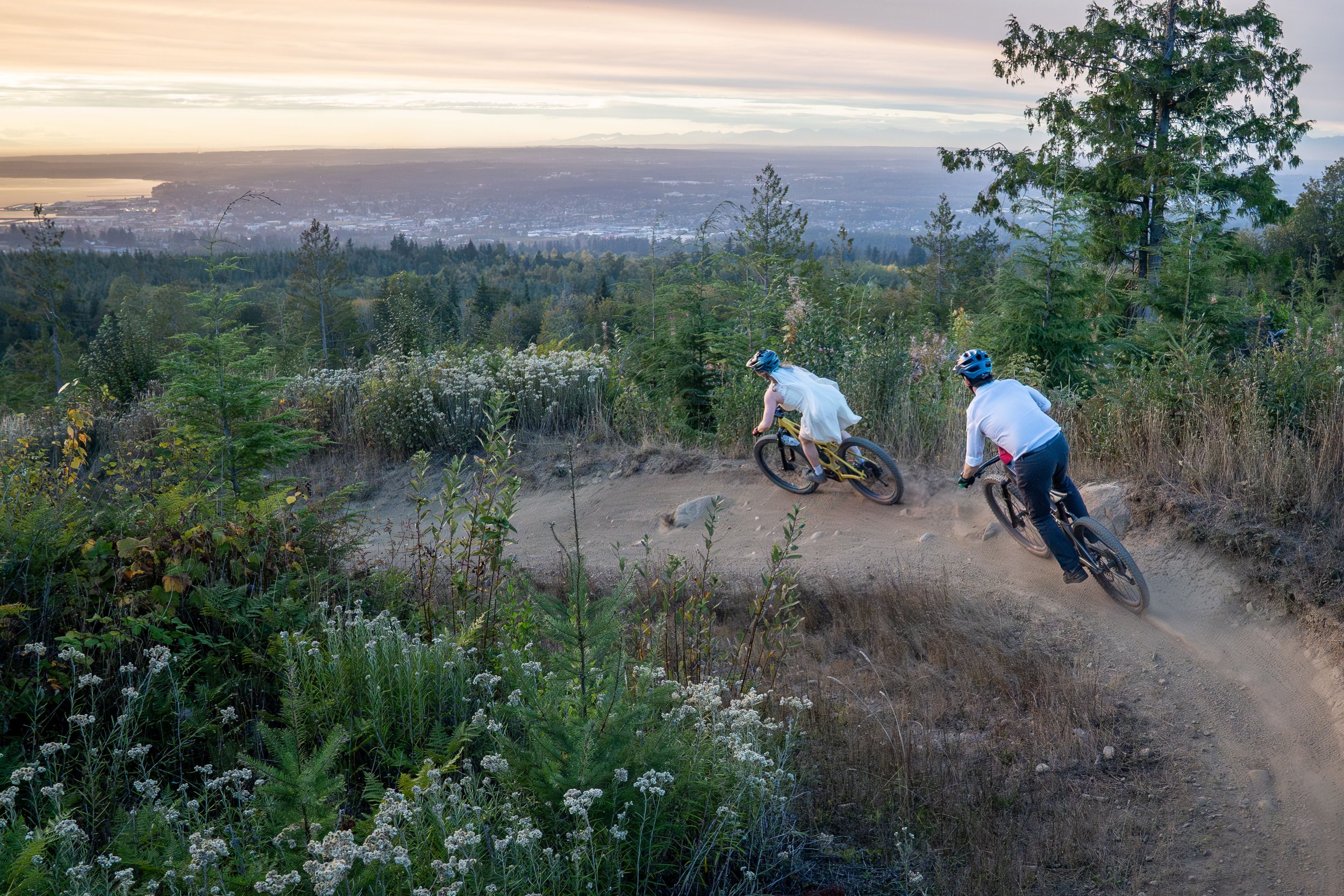 A couple in wedding clothes riding mountain bikes down SST on Galbraith Mountain at sunset during their mountain biking elopement. The riders are in the foreground, and the city of Bellingham and Bellingham Bay at sunset are in the background.