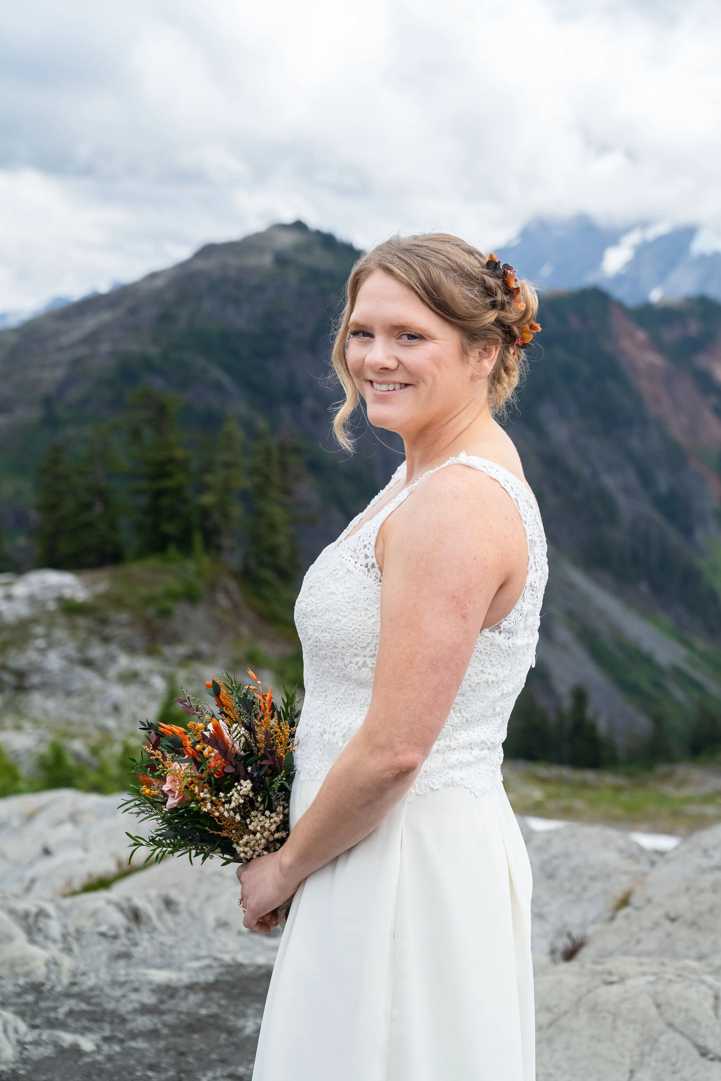 Portrait photo of a bride after her elopement in the North Cascades. She is standing in profile holding a bouquet and looking at the camera. Rocky slabs and Mount Shuksan are visible behind her.