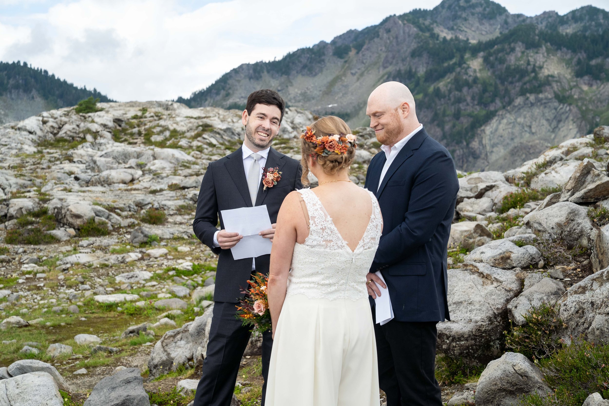 A photo of a bride, groom, and officiant holding a wedding ceremony in the mountains. The groom is smiling at the bride while reading his vows.