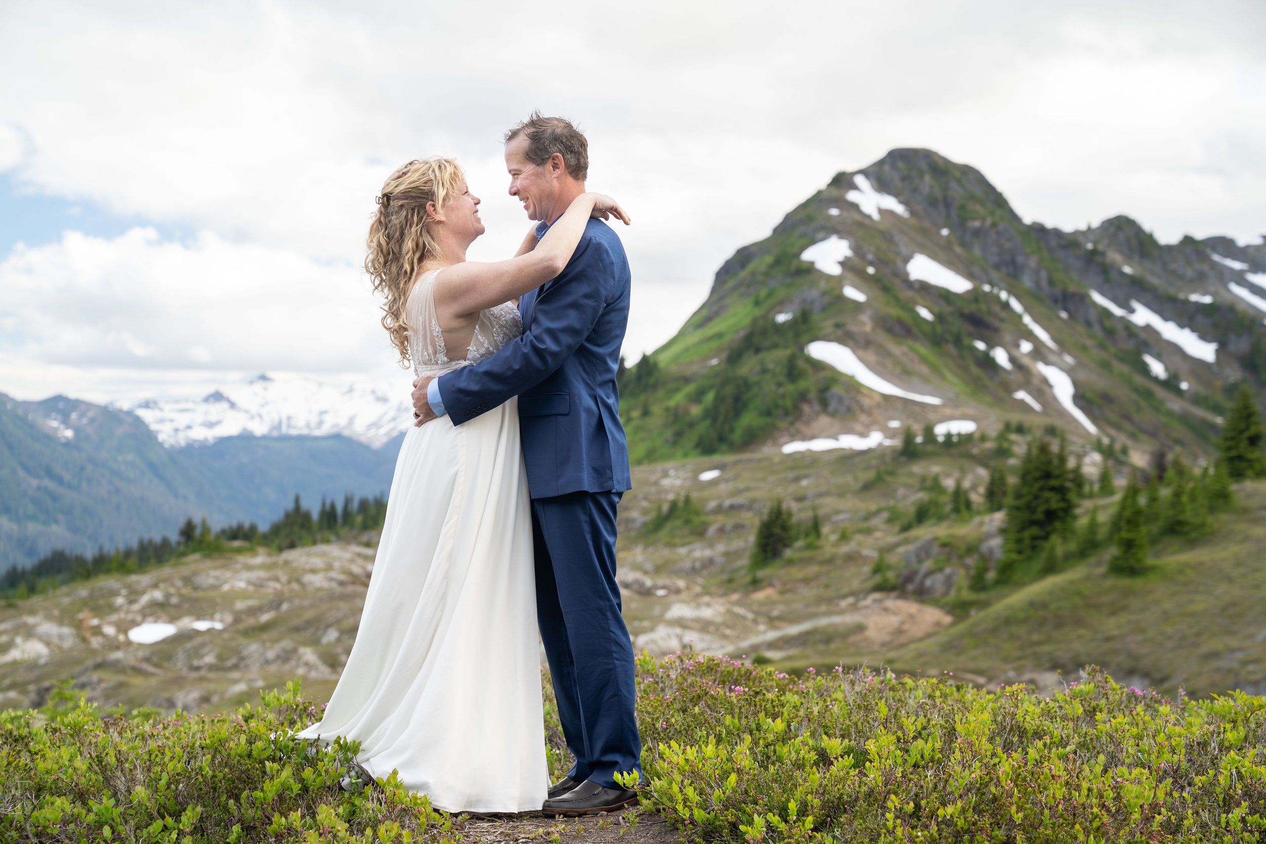 A bride in a white wedding dress and a groom in a blue suit stand together holding one another during their sunset elopement ceremony at Yellow Aster Butte in the North Cascades. They are surrounded by blooming heather and Mt. Baker is visible behind
