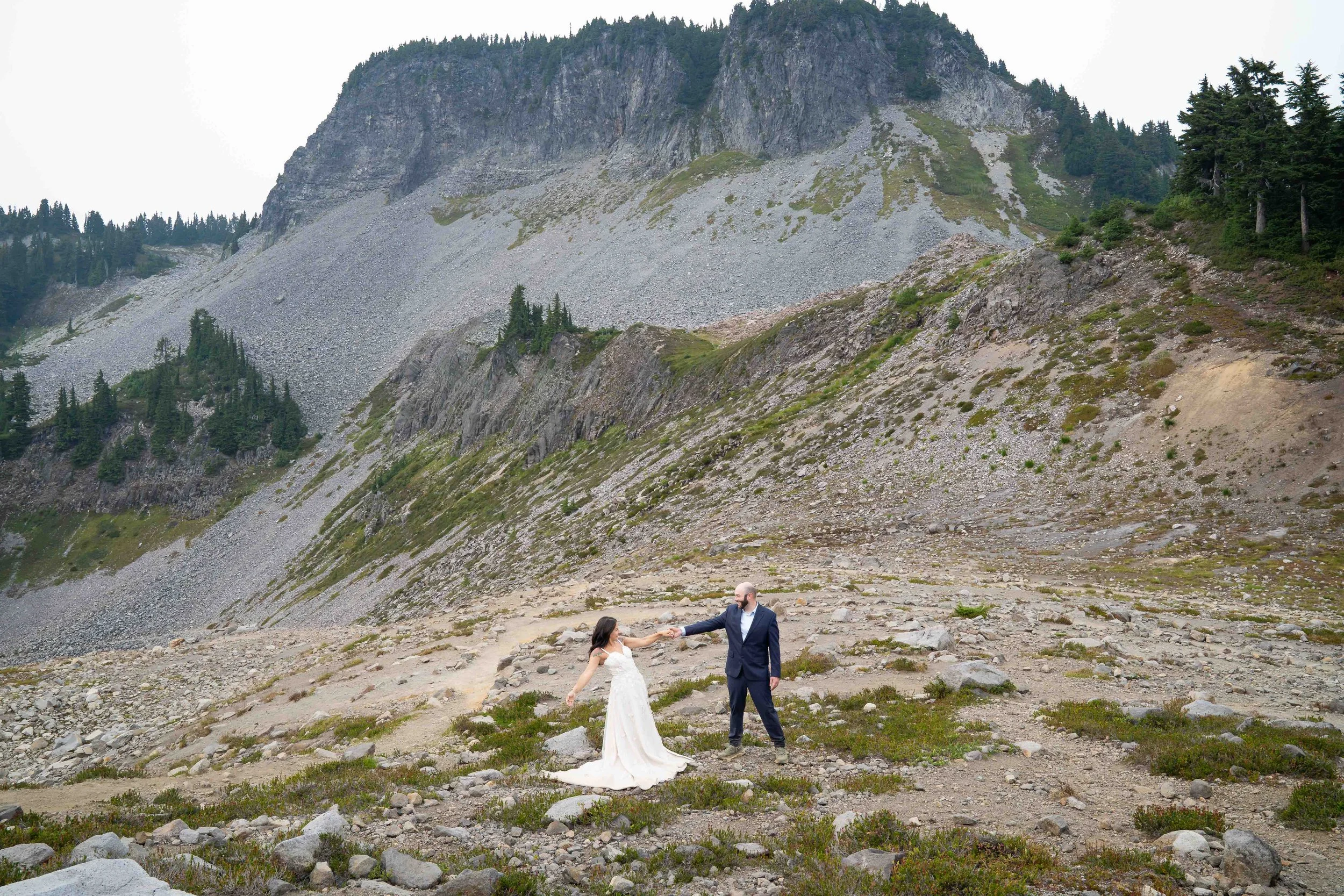 A bride and groom holding hands with their arms outstretched at the end of their first dance on Ptarmigan Ridge during their Mt. Baker elopement. They are standing in an open, rocky alpine meadow with Table Mountain behind them.