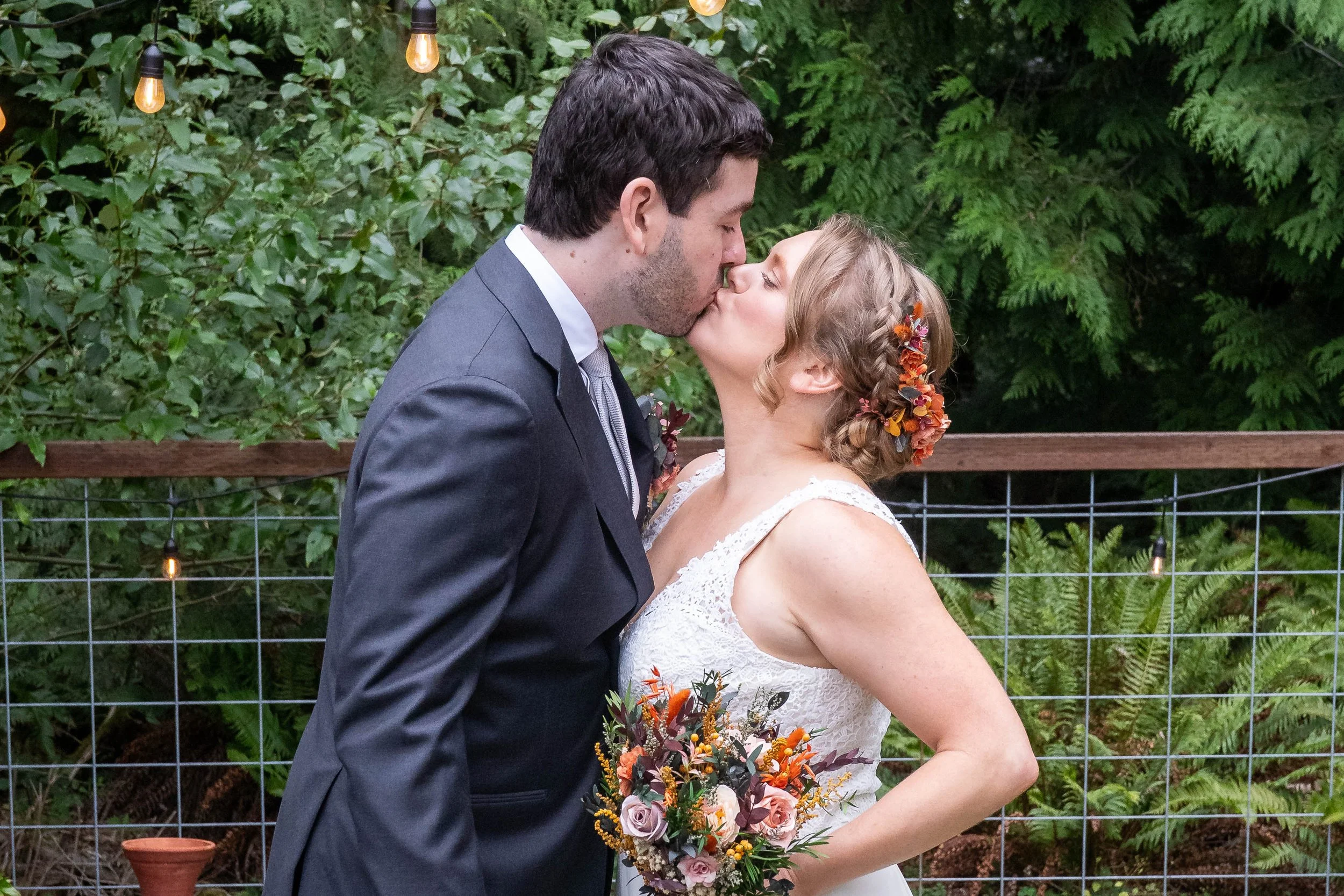 A photo of a bride and groom kissing after their first look ceremony. The bride is wearing a white wedding dress and holding a bouquet and there's a lust PNW forest in the background.