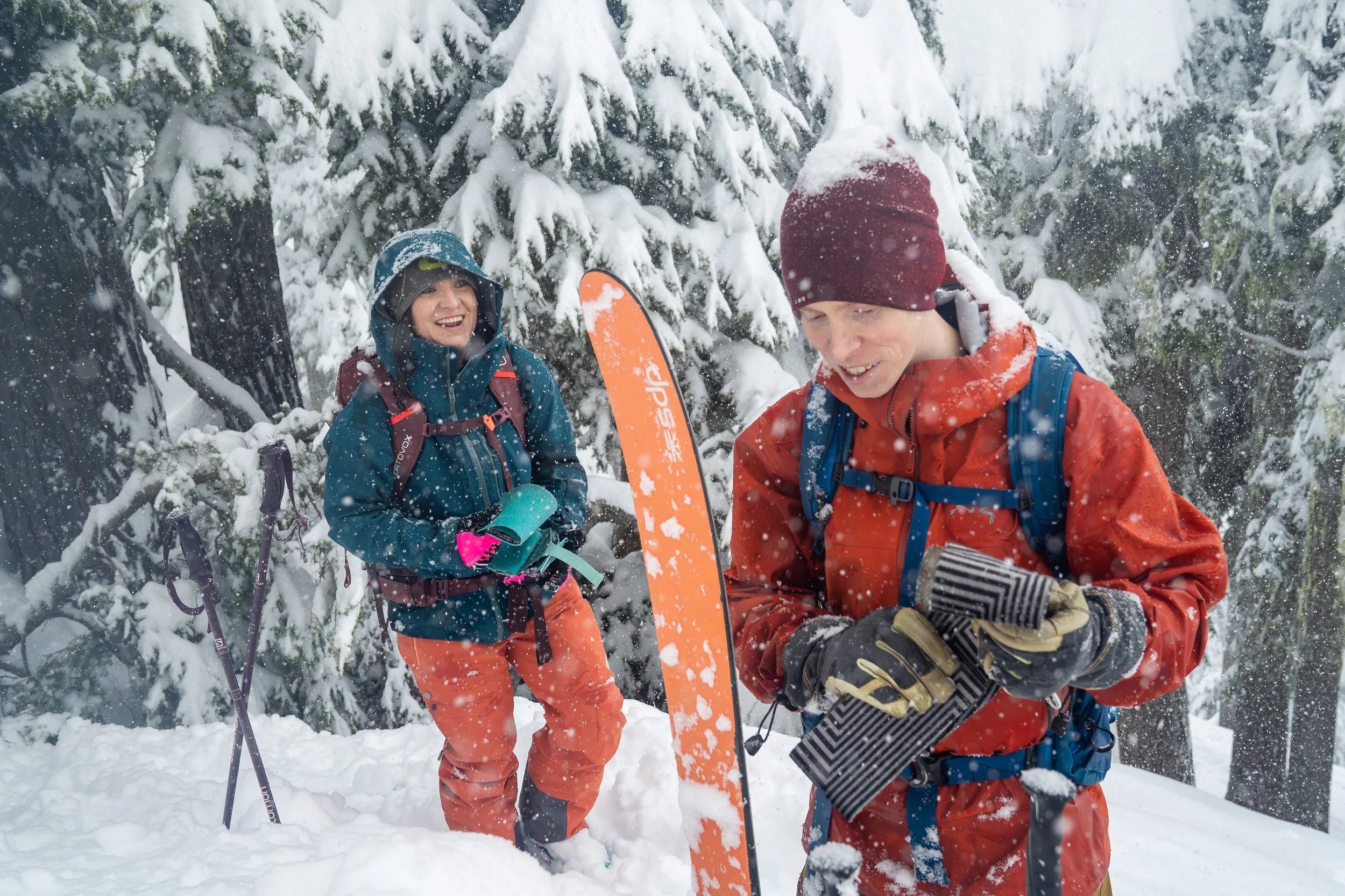 A couple with backcountry ski gear standing in the snow, each holding climbing skins, as they get ready to transition from skinning to skiing.