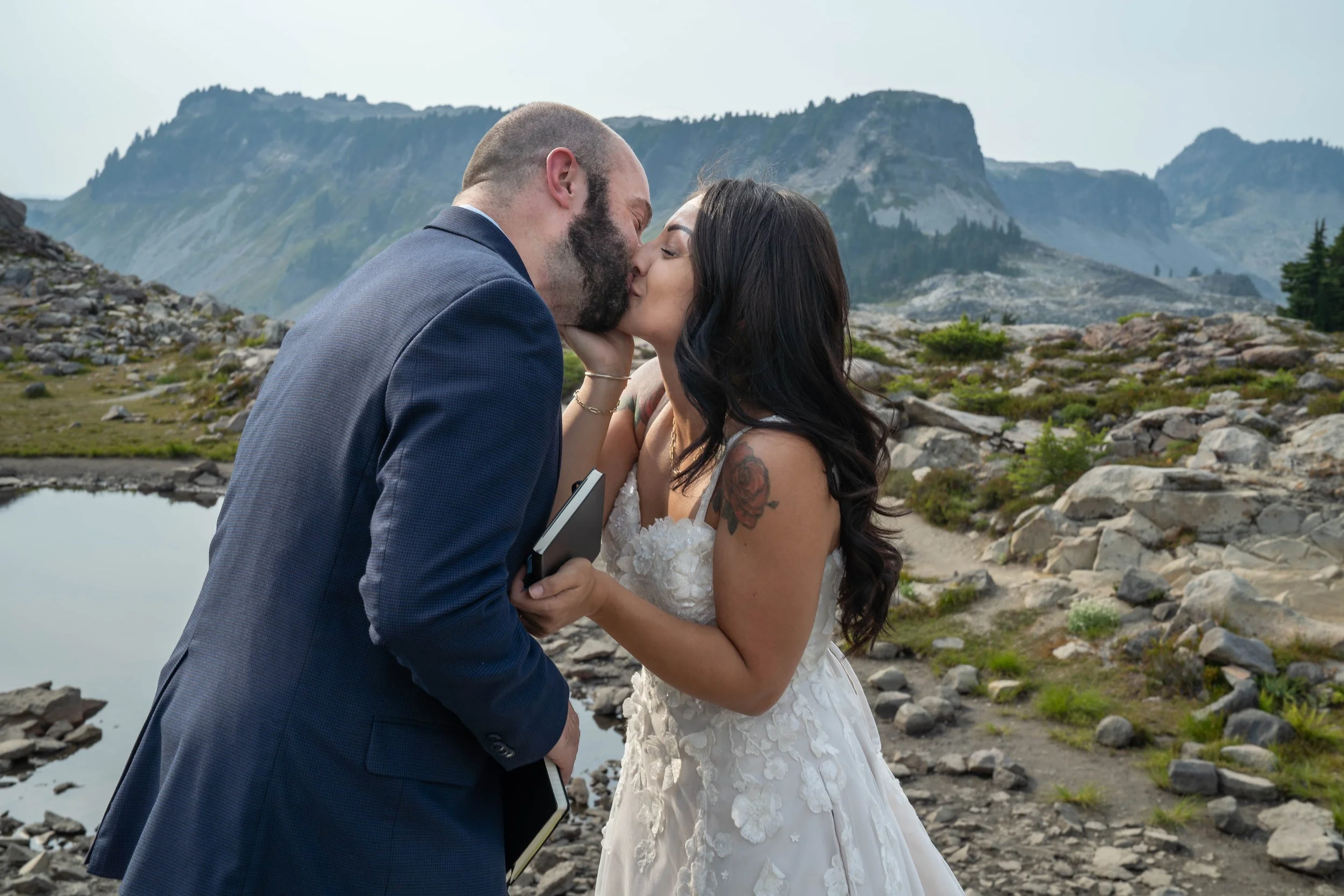 A bride and groom share their first kiss during their Artist Point elopement ceremony. They are standing in a rocky meadow with an alpine lake and Table Mountain visible behind them.
