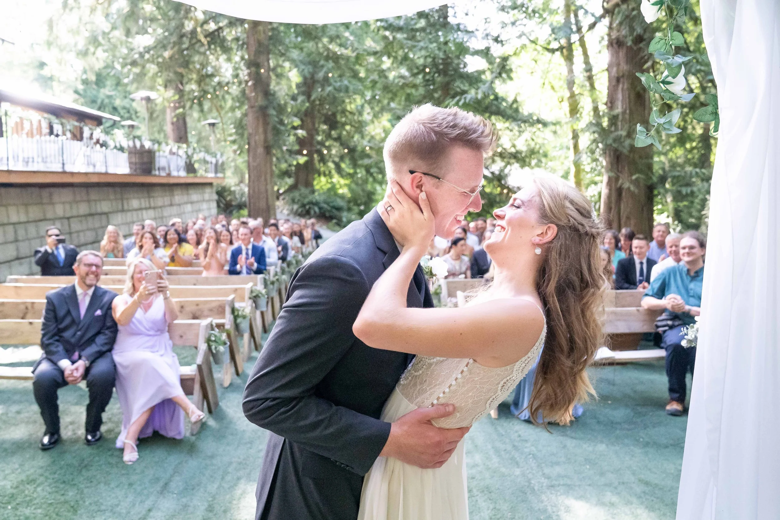 Photo of a bride and groom's first kiss during a forest wedding ceremony at Creekside Celebrations in Skagit County, Washington. The photo is taken from behind the altar and the wedding guests are visible behind the couple.