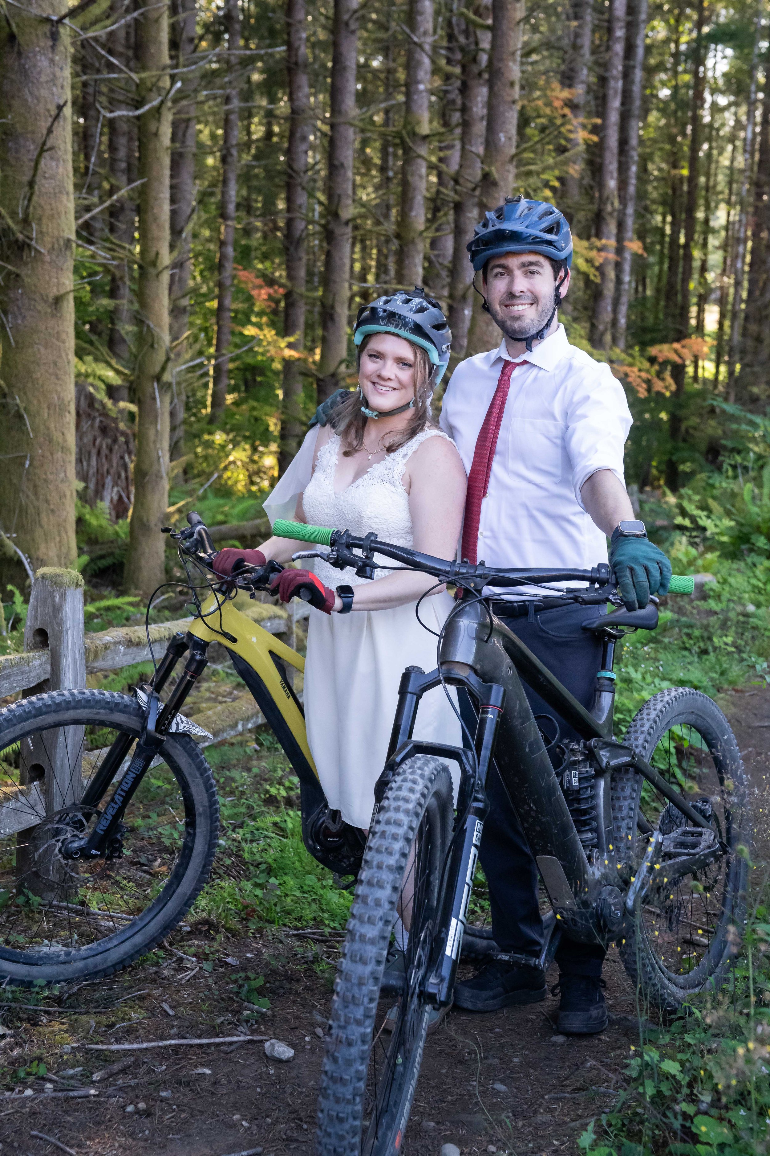A bride and groom standing together with their bikes during a mountain bike elopement on Galbraith Mountain in Bellingham, Washington. The couple is wearing full wedding clothes plus helmets and bike gloves.