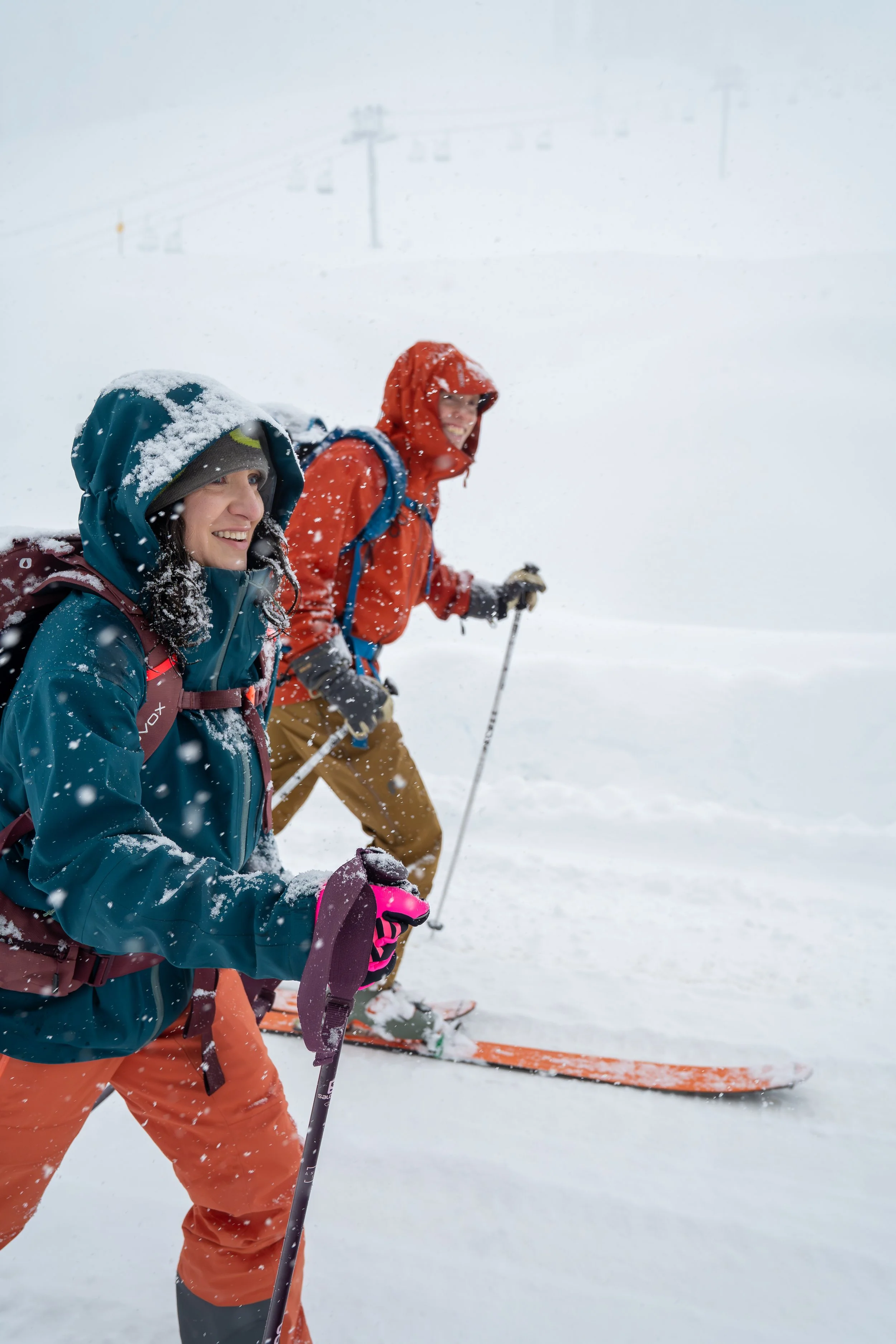 A couple backcountry skiing up a snowy road, with the Alpental ski lift in the fog in the background. It's snowing and the skiers have their hoods up, but they're smiling.
