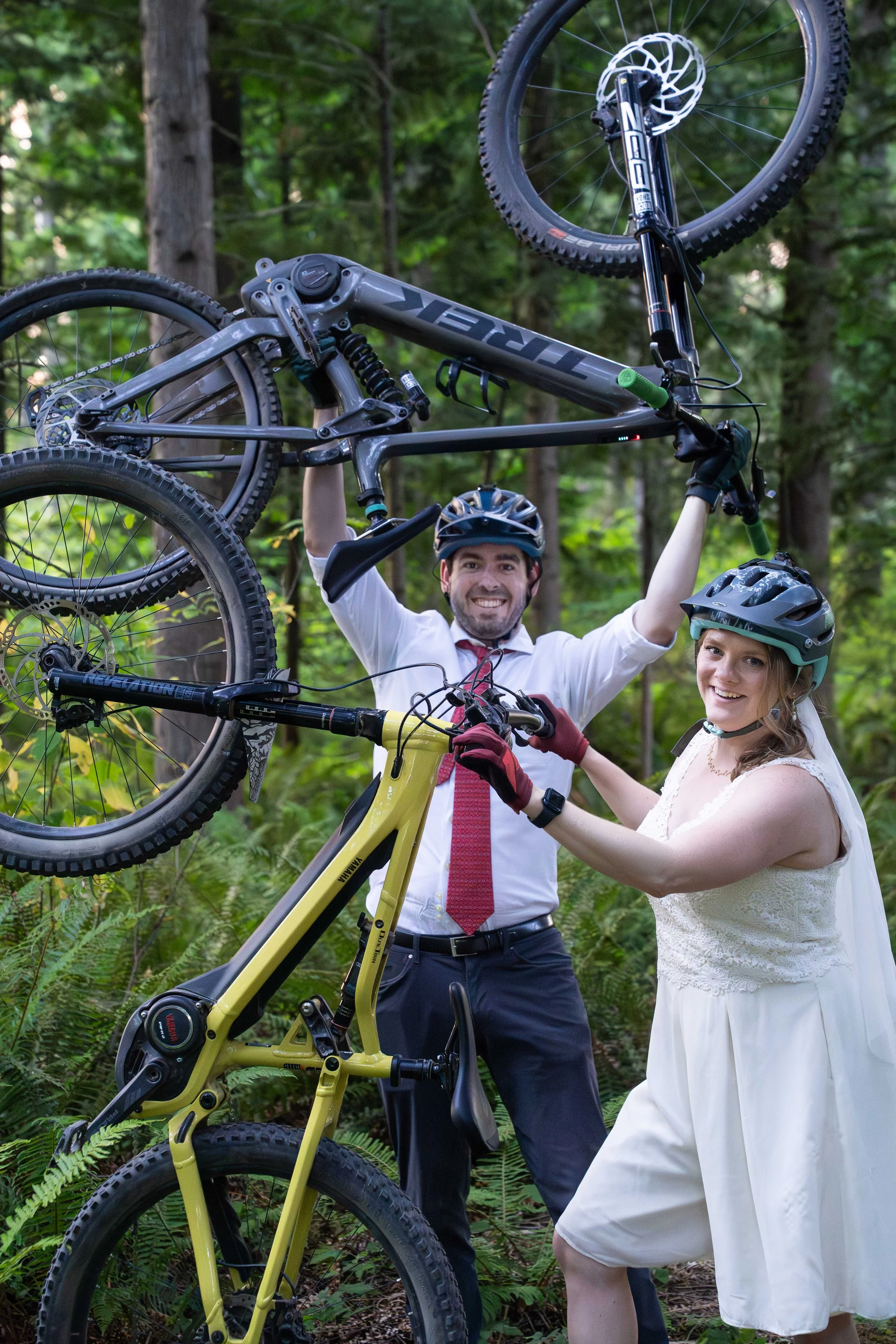 A bride and groom holding up their mountain bikes over their heads while wearing full wedding clothes during a mountain biking elopement photoshoot on Galbraith Mountain, in Bellingham, Washington. They are on singletrack surrounded by ferns.