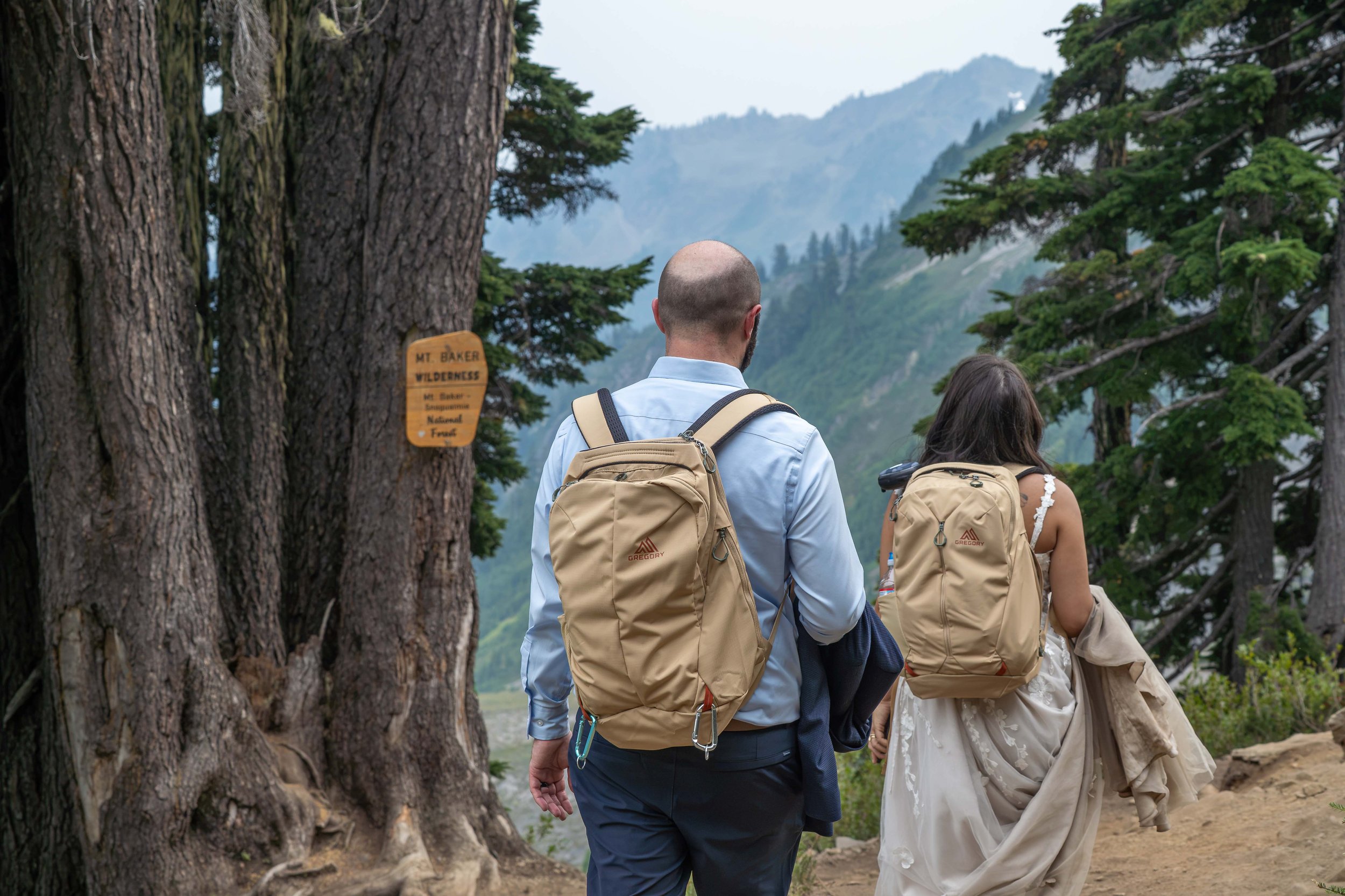 A bride and groom in wedding clothes and matching backpacks hike the Ptarmigan Ridge trail during their Mount Baker elopement. They're hiking among large fir trees with mountains visible through the trees.