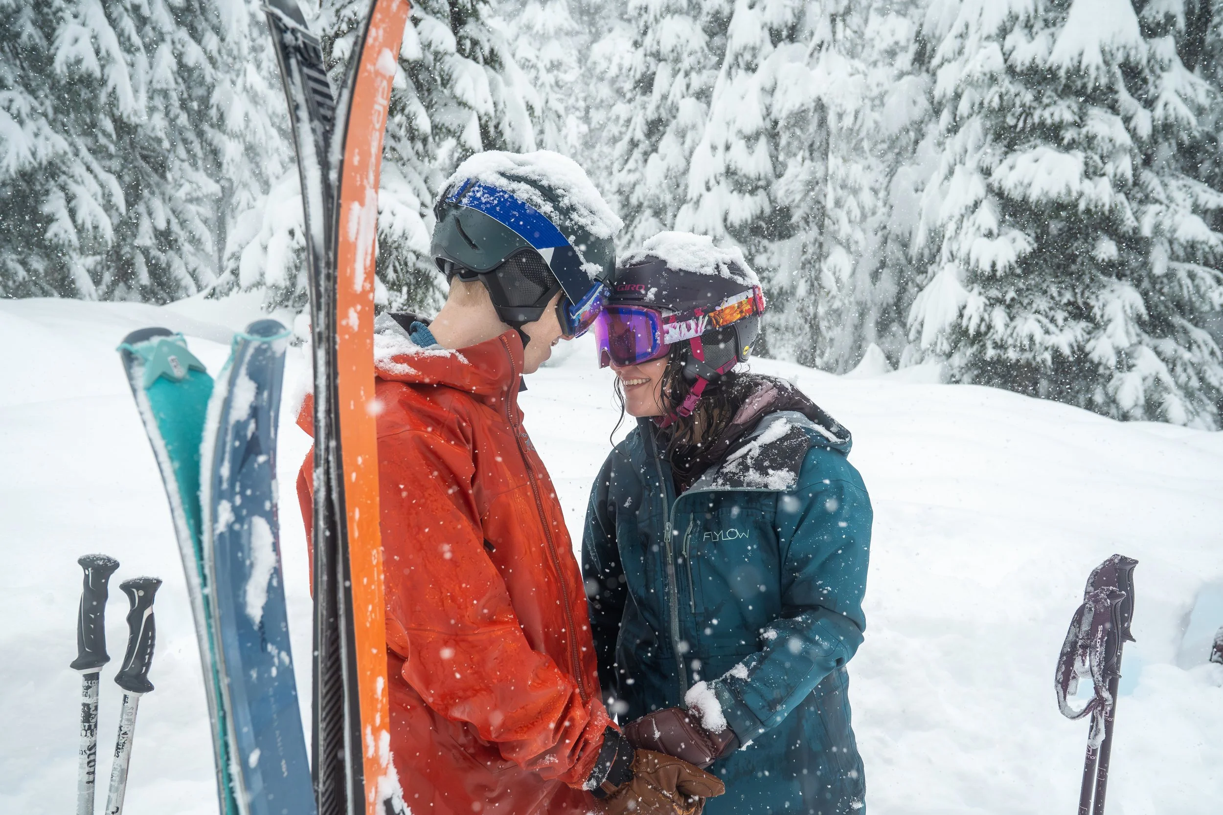 A photo of a man and woman in ski gear, including ski goggles and helmets, standing together in the snow and pressing their helmets together in an intimate pose. Their skis and poles are visible in the foreground and the forest behind them is snowy.
