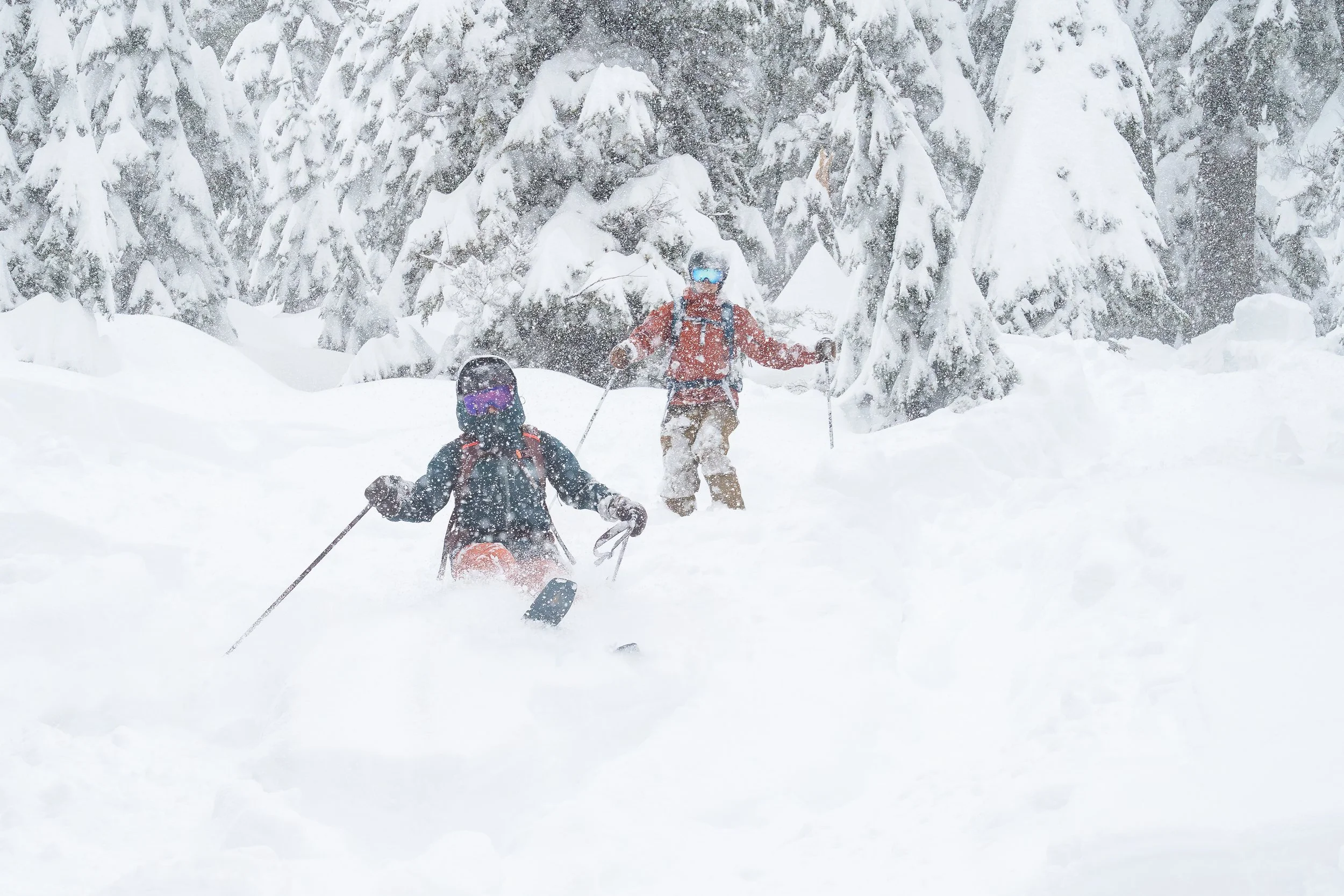 A couple backcountry skiing in deep powder in the North Cascades during a winter backcountry ski elopement. They're wearing ski jackets and ski goggles and it's snowing heavily as they ski down.