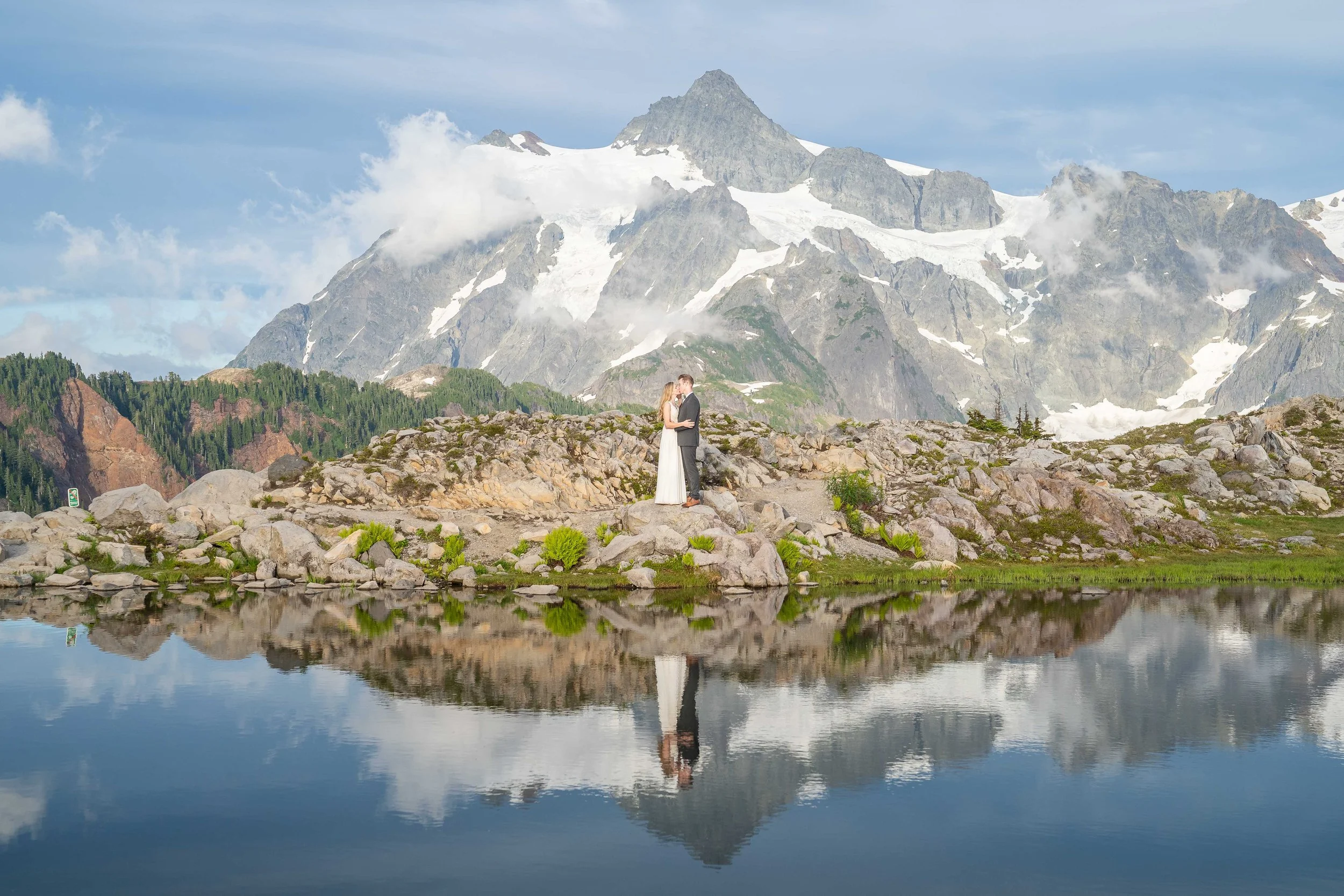A photo of a bride and groom kissing in front of an alpine lake and Mt. Shuksan during an elopement at Artist Point. The couple and Mt. Shuksan are reflected in the waters of the lake.