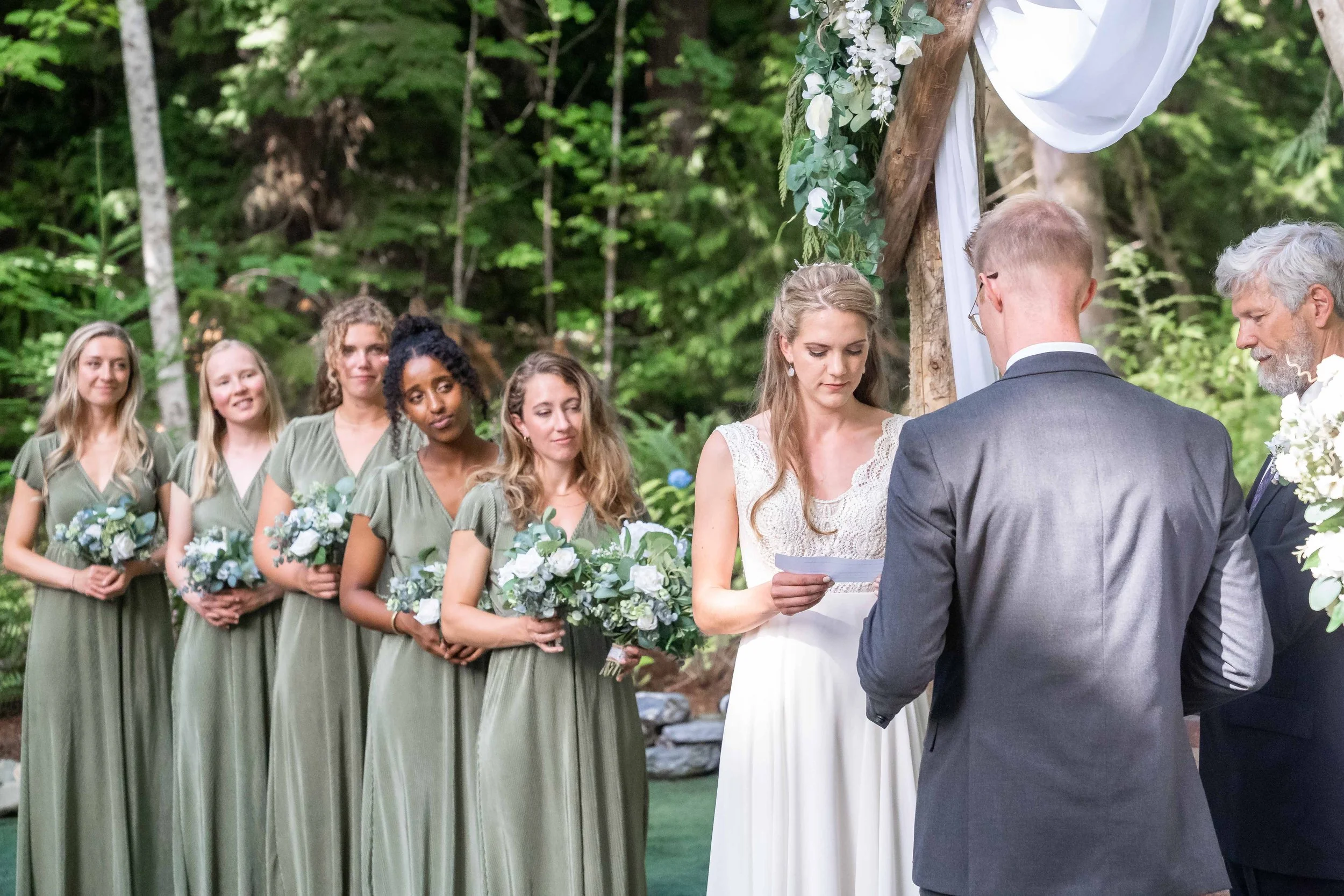 Photo of a wedding ceremony in the woods at Creekside Celebrations in Skagit County, Washington. The bride is reading her vows while the groom and her bridesmaids look on.