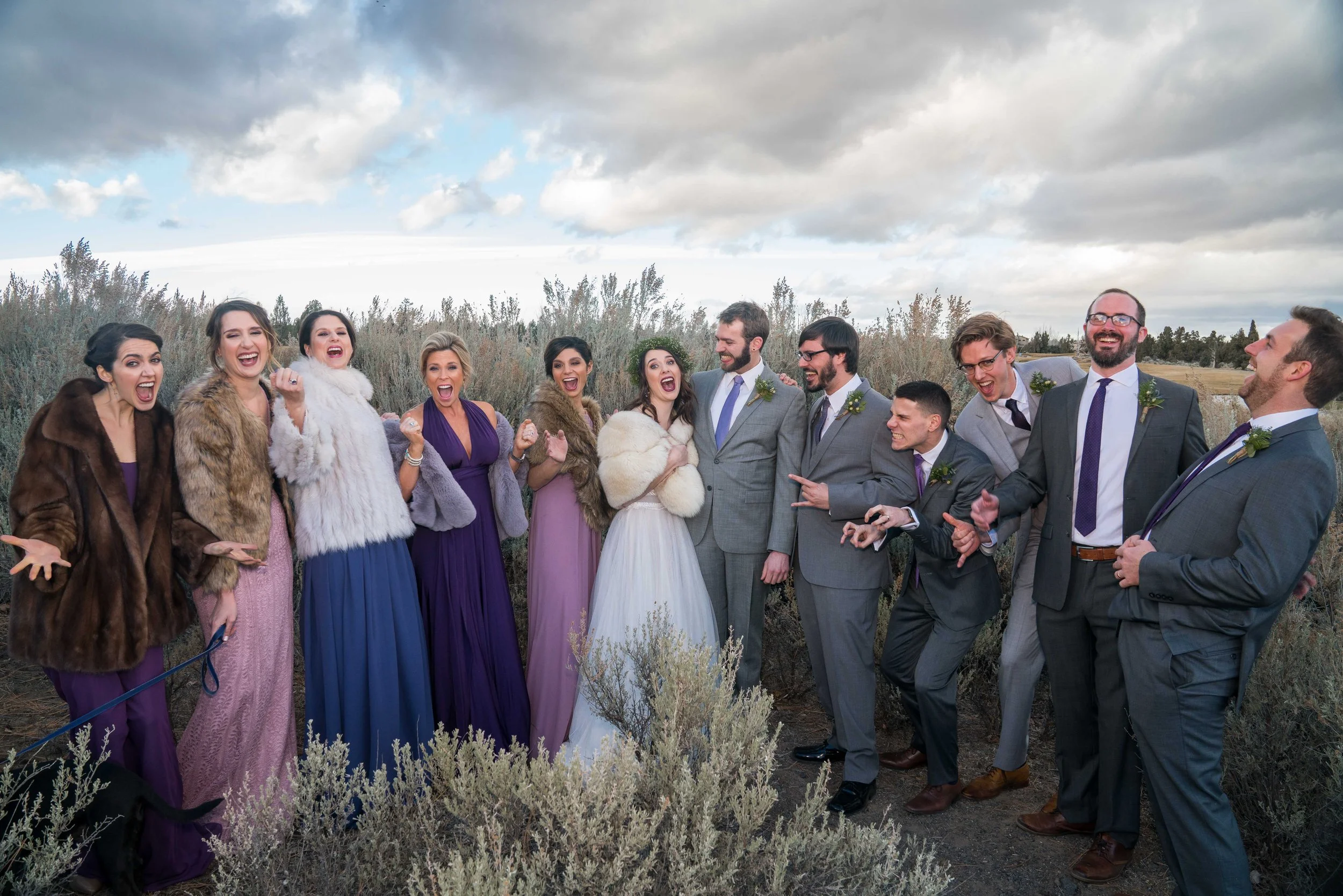 A photo of a wedding party surrounding the bride and groom. The groomsmen are all on one side and the bridesmaids are all on the other, and they're looking excited and yelling towards the camera.