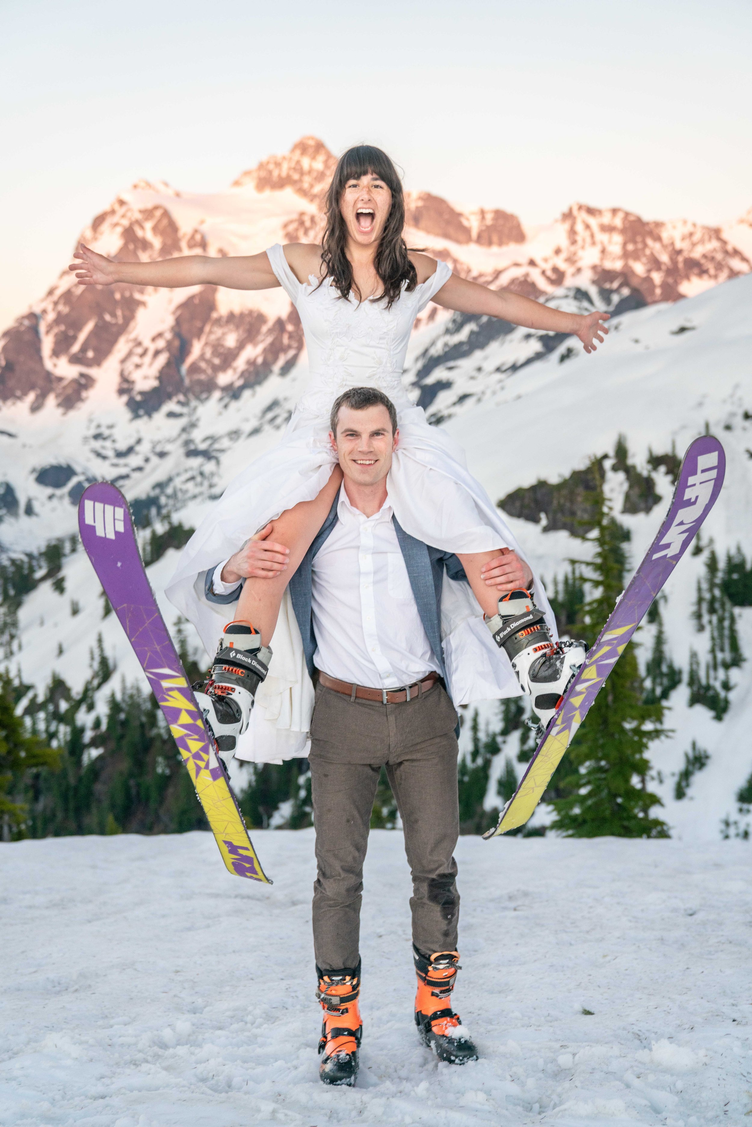 Photo of a woman in a wedding dress and with skis on sitting on her husband's shoulders with Mount Shuksan in the background.