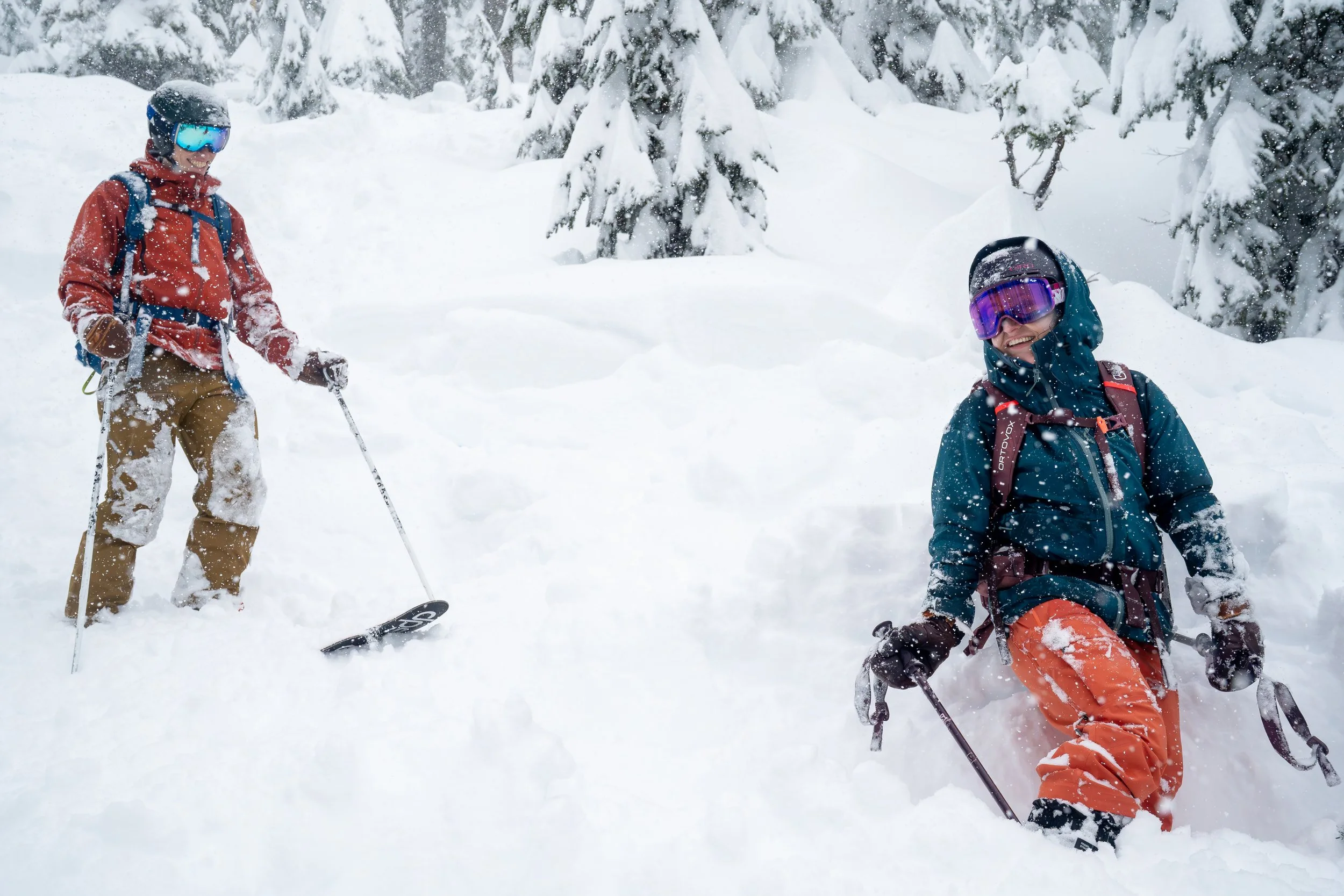 Photo of a couple on skis in deep snow in the backcountry around Snoqualmie Pass. The woman is looking back and smiling at the man and they're both covered in fresh snow.