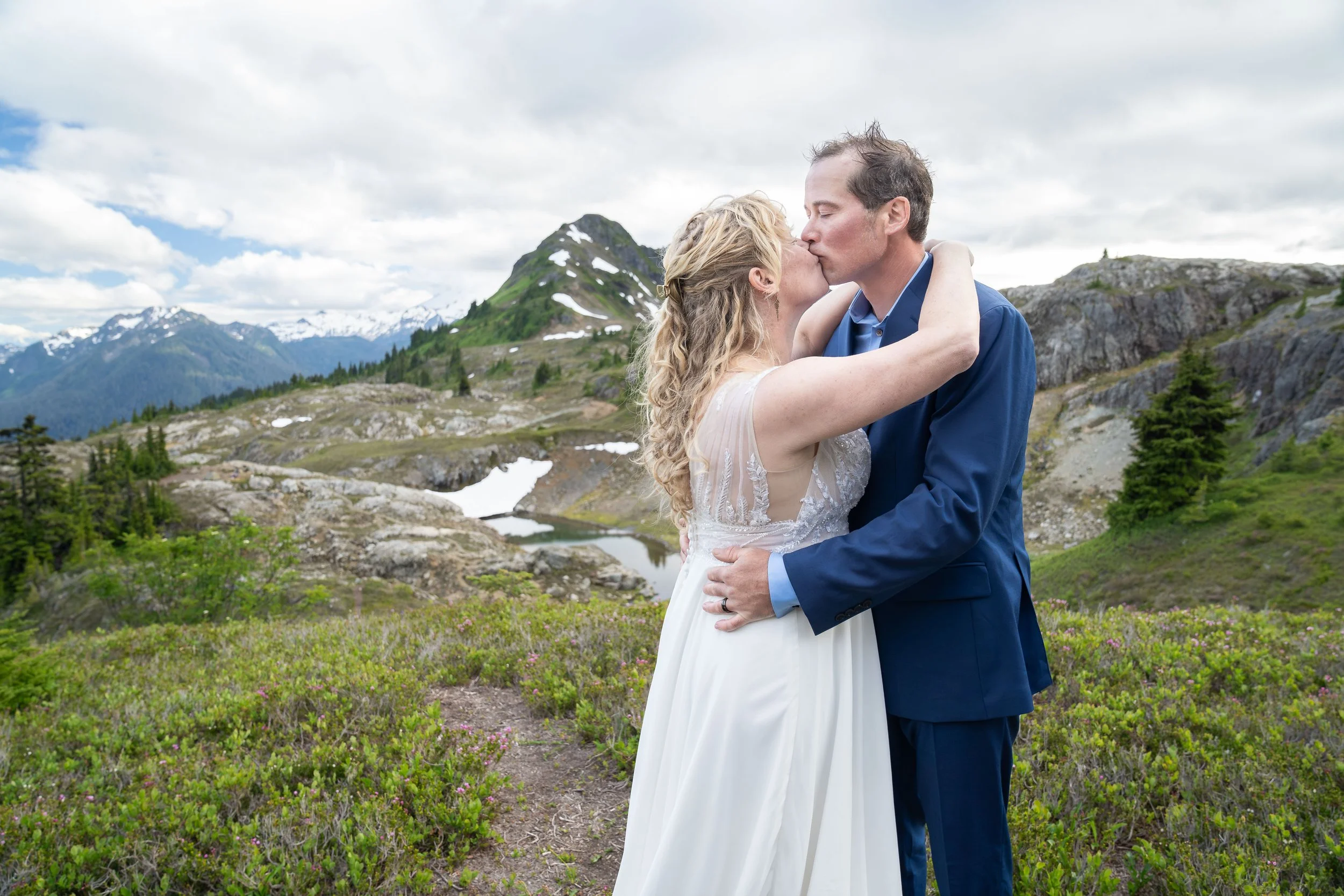 A bride and groom in wedding clothes kissing in a subalpine meadow in the North Cascades, Washington. They are near the High Divide trail and Keep Kool Butte and Mt. Baker are visible in the background.