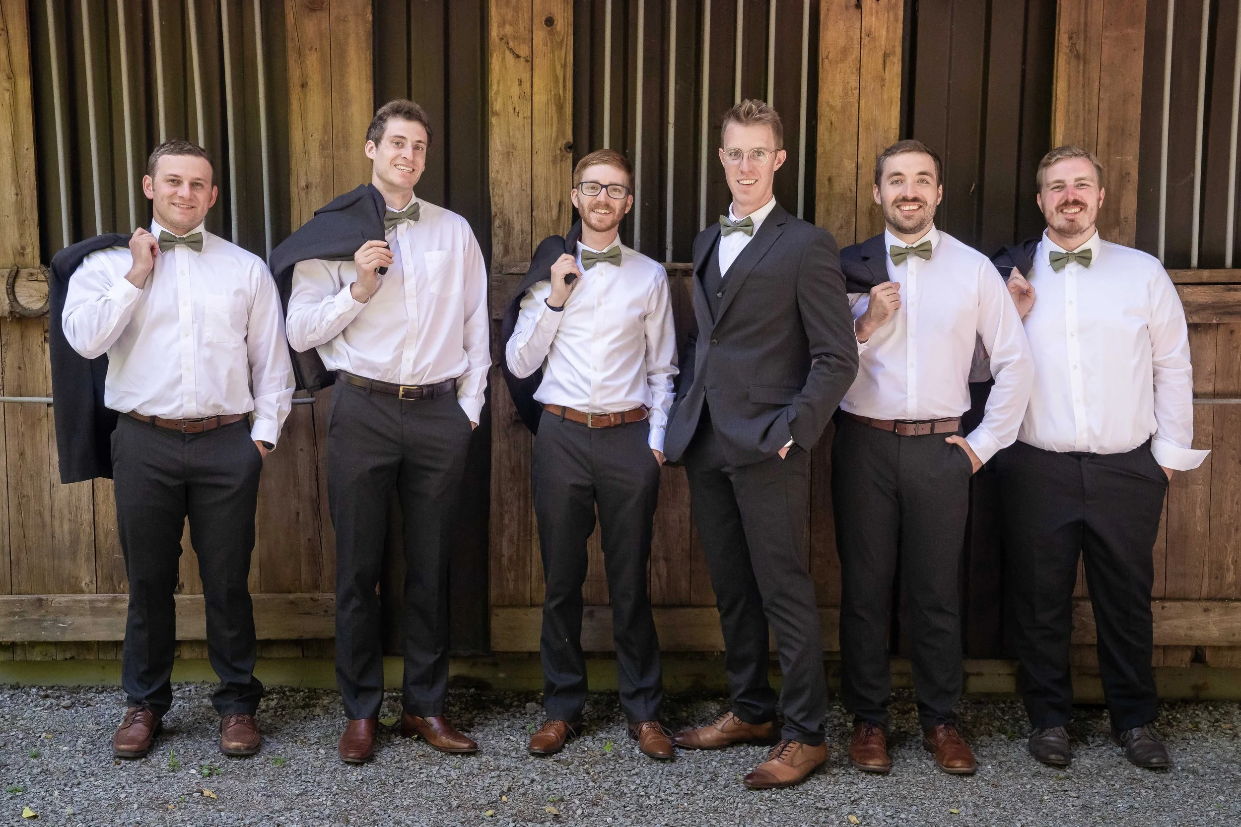 A photo of a groom and his groomsmen at a wedding in Skagit County, Washington. The men are standing in front of a wooden stable. The groom has a black suitjacket on while the groomsmen have tossed their jackets over their shoulders.