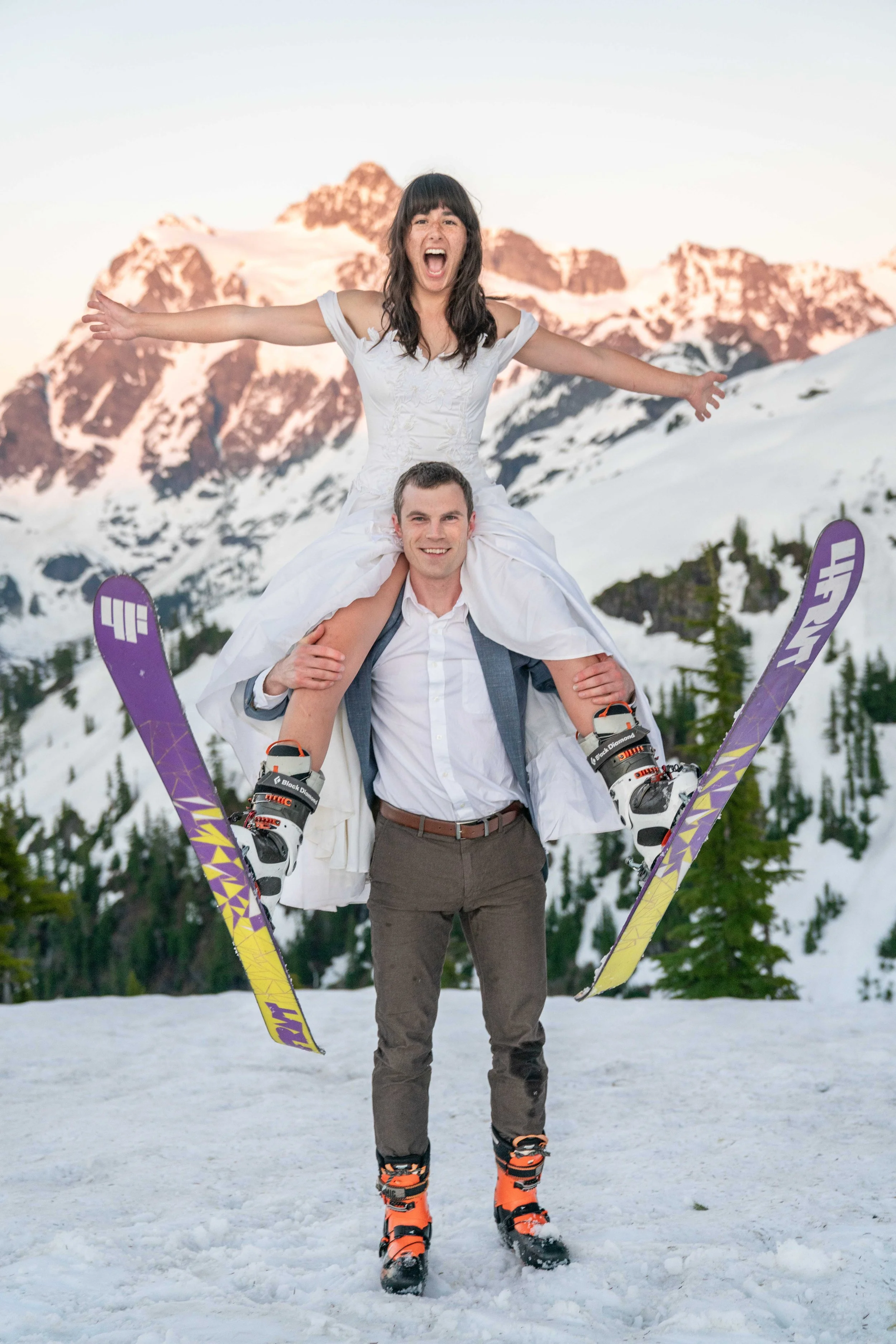 A photo of a bride in a wedding dress and with skis on sitting on her husband's shoulders and spreading her skis. He's wearing a suit and ski boots. Mount Shuksan at sunset is behind them.