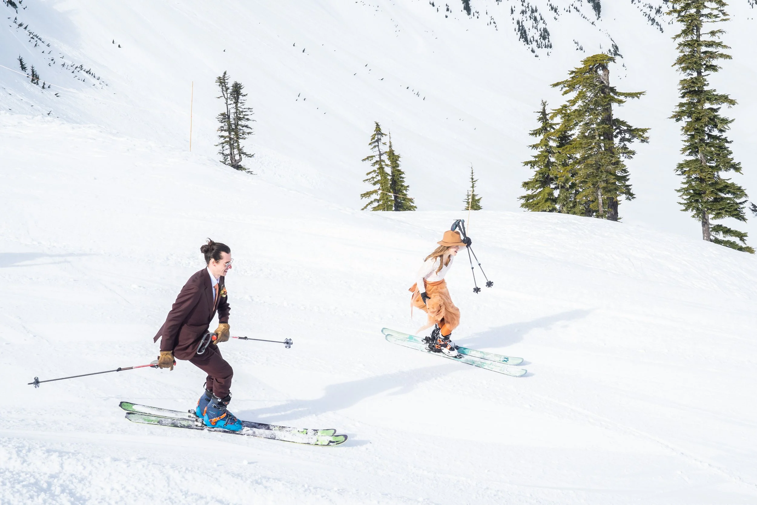 A photo of a couple skiing together in the Baker Backcountry, Washington. The couple is in wedding clothes with the groom wearing a dark brown suit and the bride wearing an orange wedding dress.