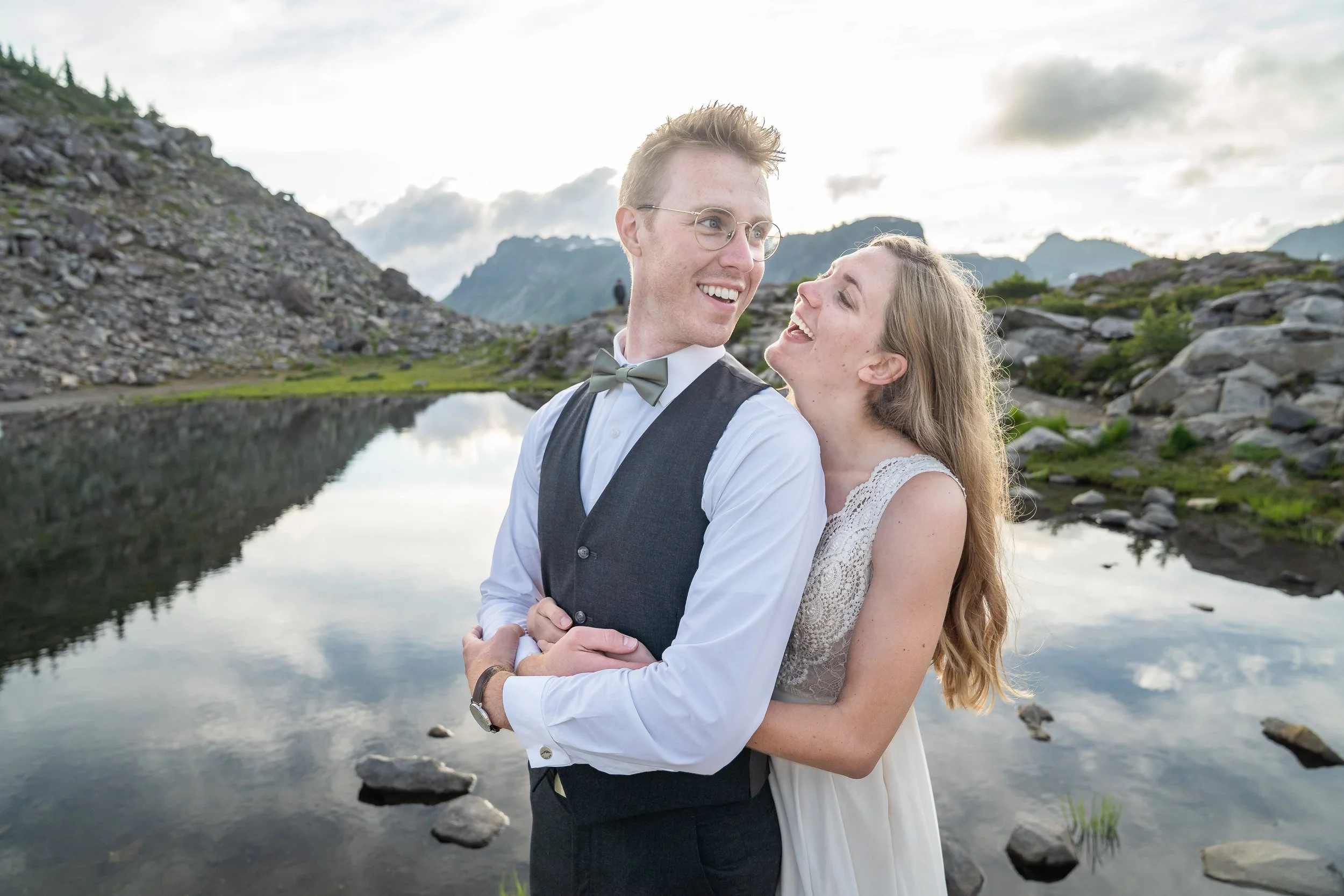 Photo of a couple in wedding clothes smiling at each other with an alpine lake and sunset in the background.