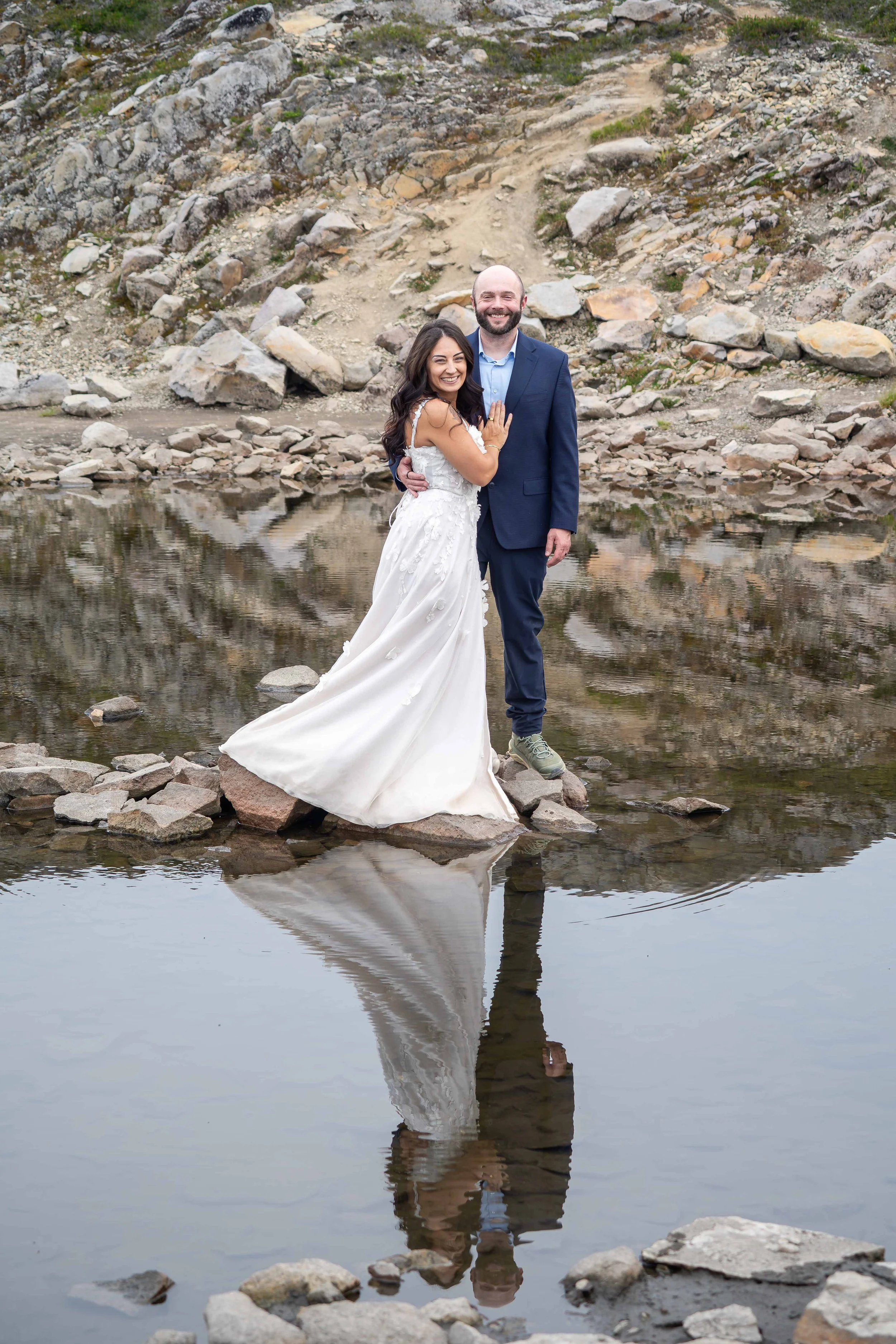 A bride and groom in wedding clothes are standing on rocks in the middle of an alpine lake and smiling at the camera during their Artist Point elopement. Their reflection is mirrored in the lake.