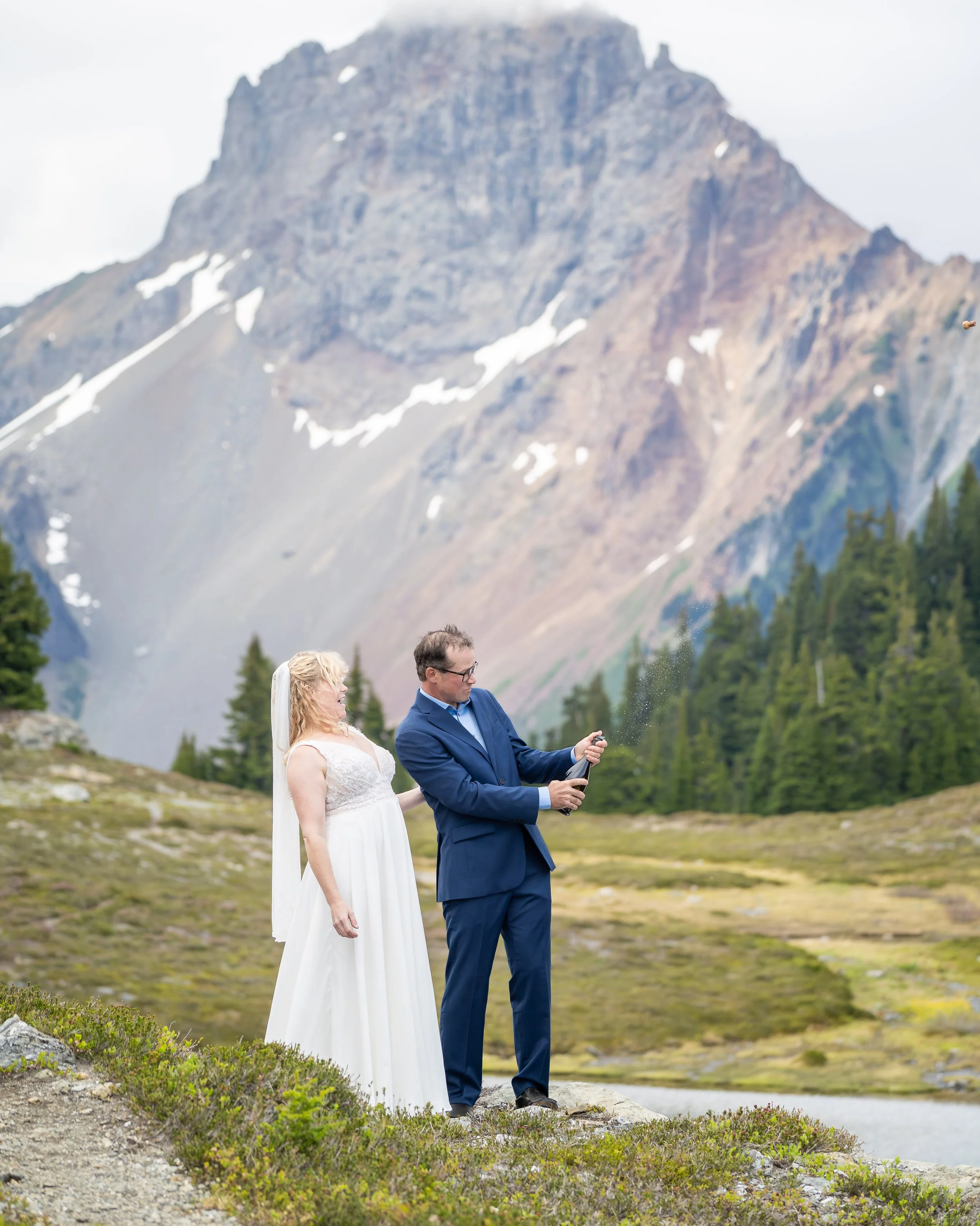 A bride and groom pop champagne during their North Cascades elopement at Yellow Aster Butte. They are standing in an alpine meadow next to a lake with American Border Peak behind them and the cork is visible flying into the air.