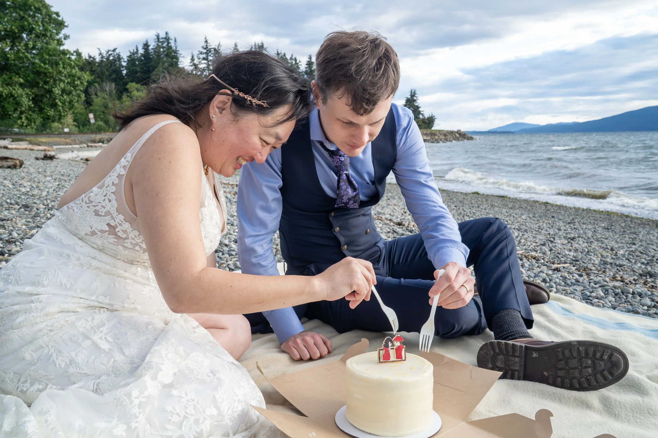 A bride and groom smile and laugh as they dig into a Pure Bliss wedding cake during a post-elopement picnic on the beach at Marine Park in Bellingham.