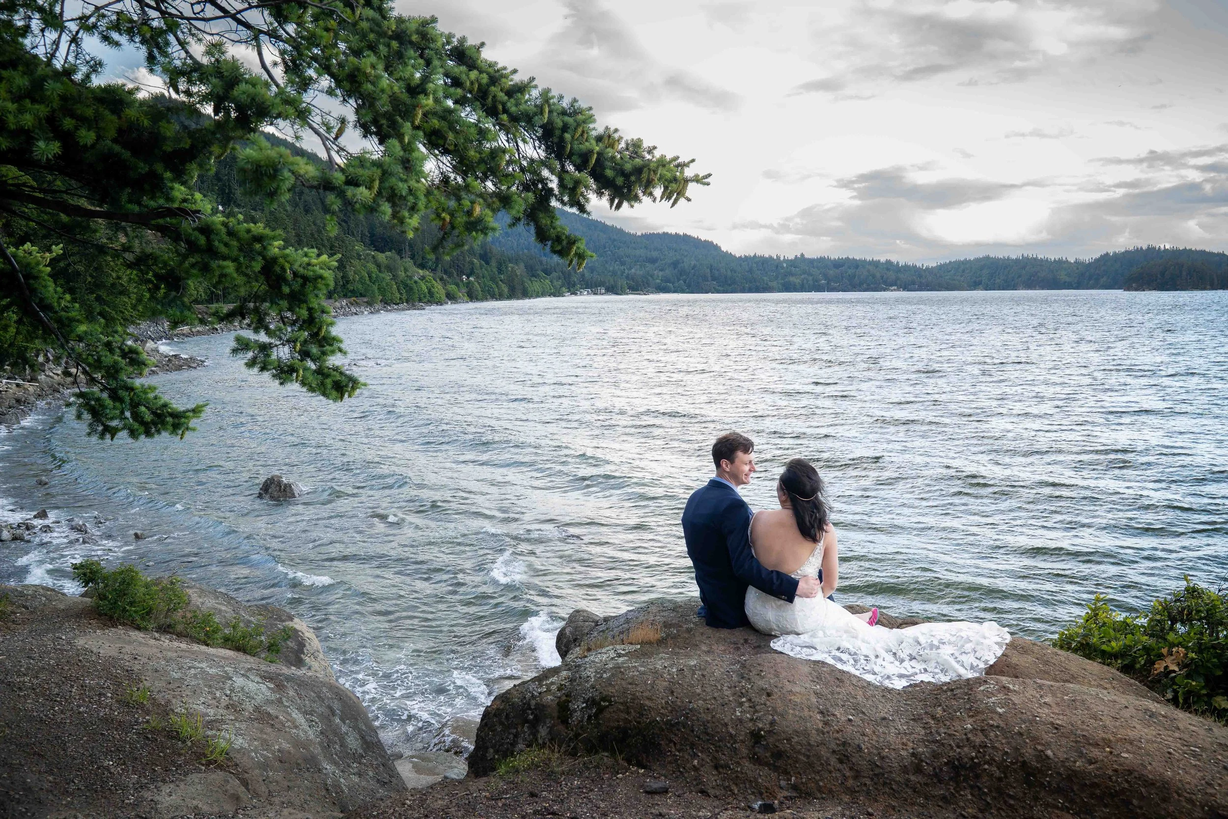 A bride and groom sit together on a rocky bluff at Teddy Bear Cove after their Bellingham, Washington elopement. They are smiling at each other and looking out over the water at sunset. The photo is framed by an evergreen tree in the foreground.
