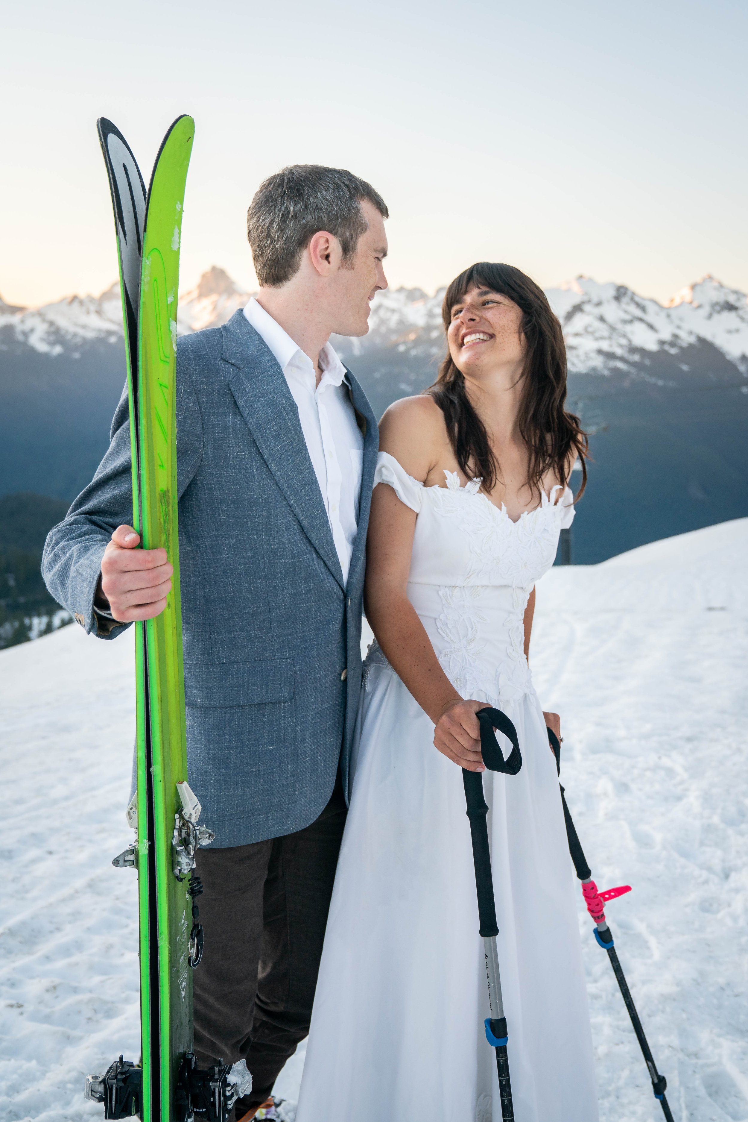 A bride and groom standing together and holding skis during a spring backcountry ski elopement neat Artist Point and the Mt. Baker Ski Area. It's sunset and the snowy peaks of the North Cascades are visible behind them.