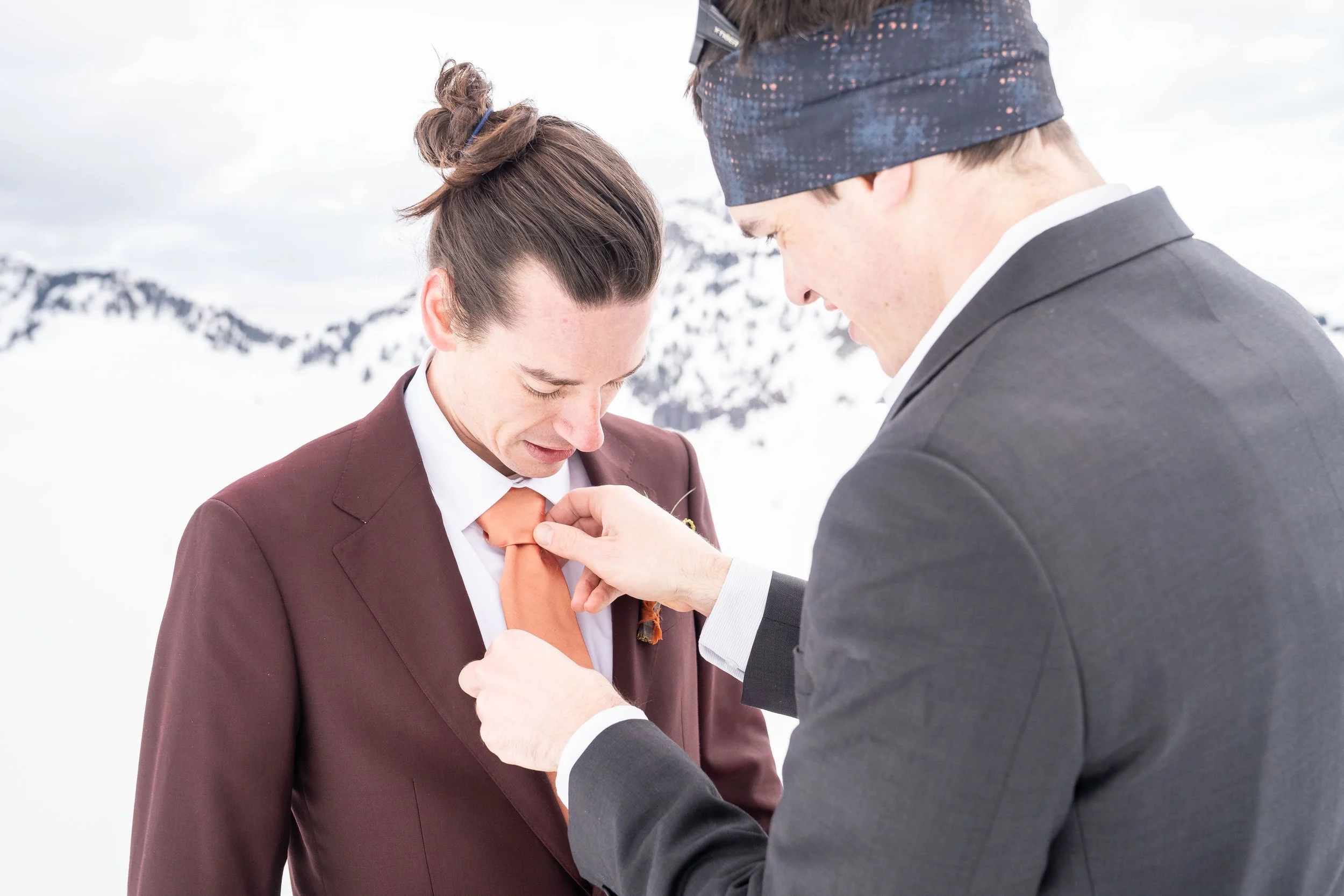 A groom and his best man standing in the snow with mountains in the background. The best man is adjusting the groom's orange tie.