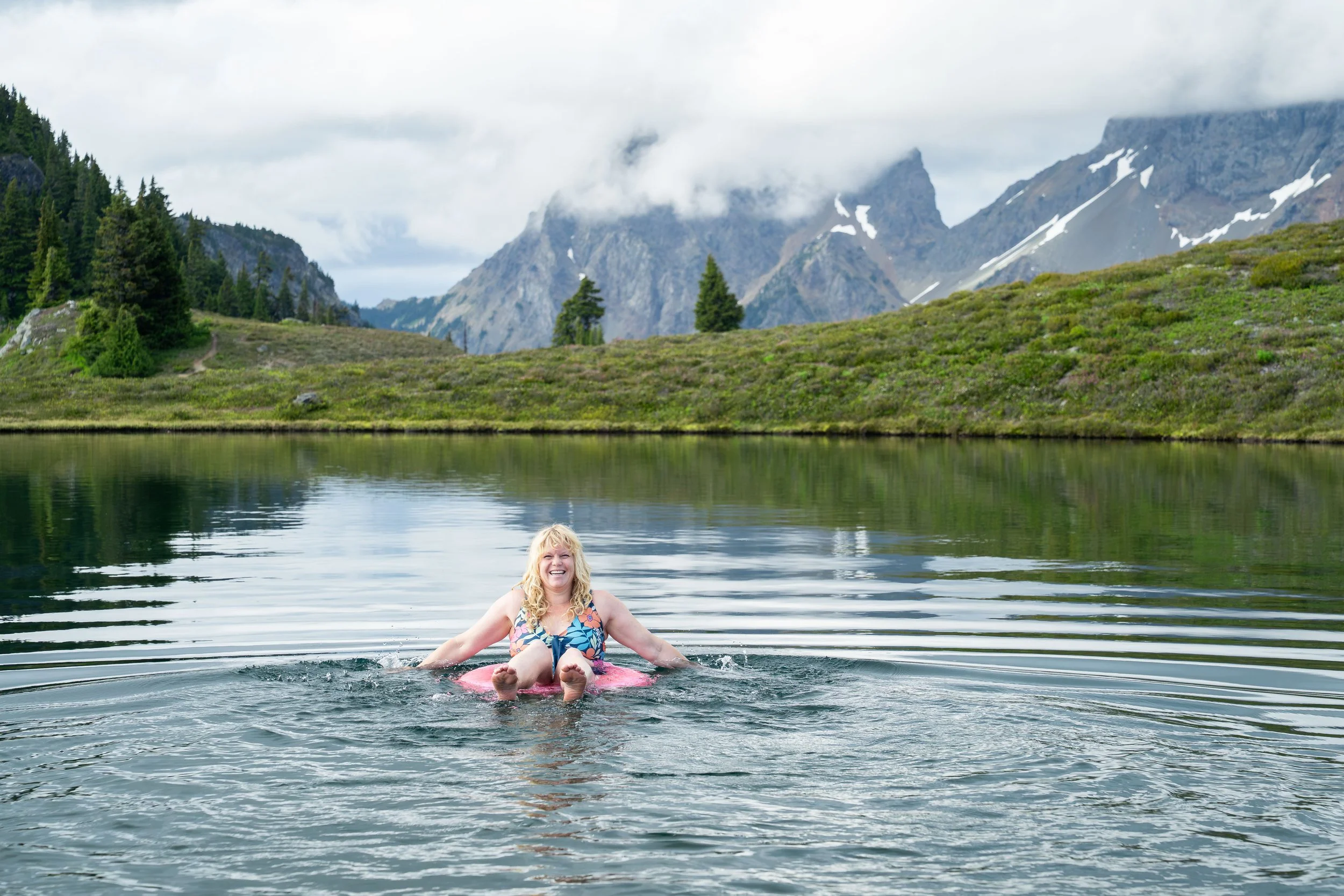 A bride floating on a bright pink innertube in an alpine lake during her elopement in the North Cascades, with wildflower meadows and American Border Peak visible in the background.