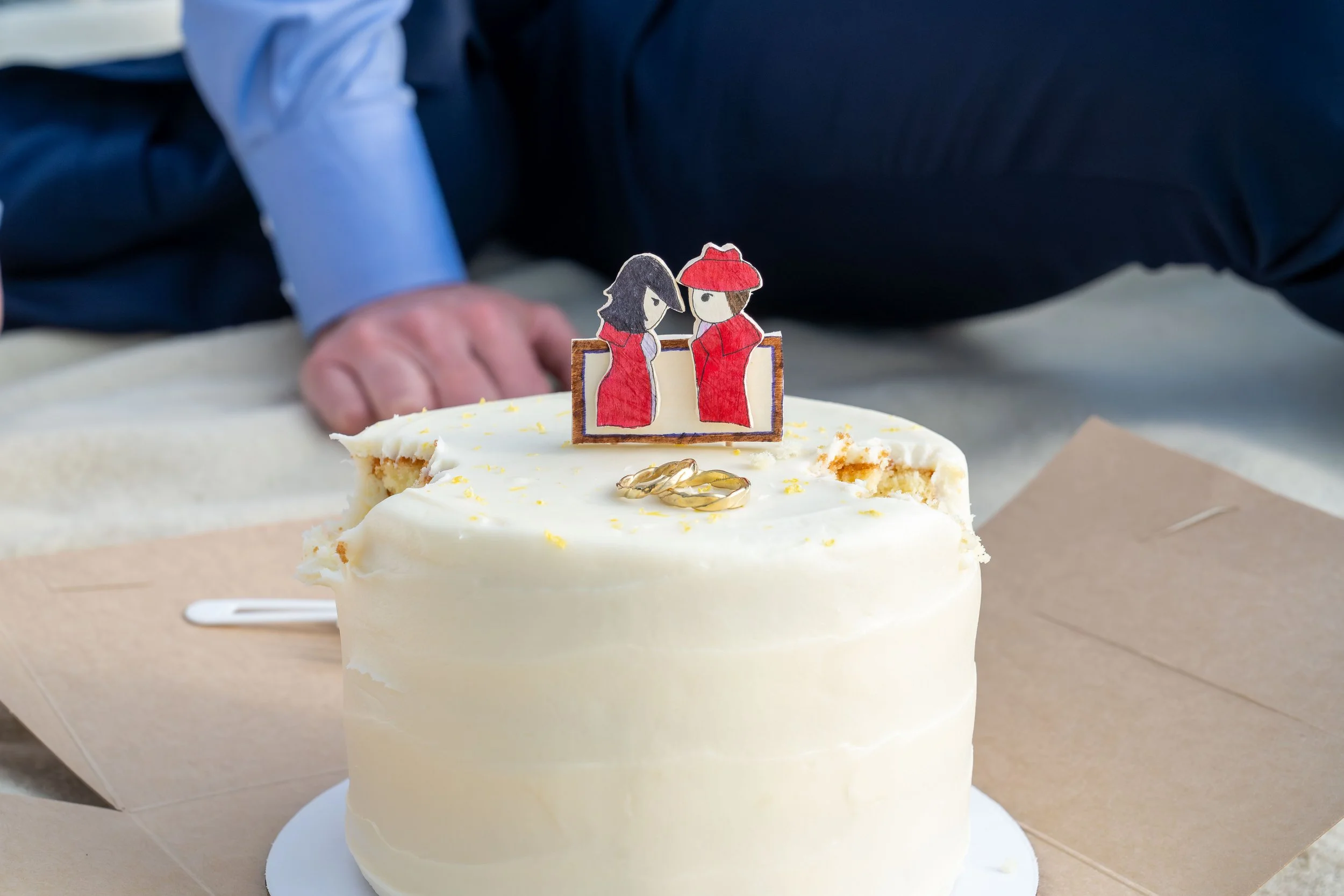 A close-up photo of a Pure Bliss wedding cake with two bites taken out of it. The couple's wedding rings are lying on top of the cake and they have a handmade cake topper that depicts the bride and groom.
