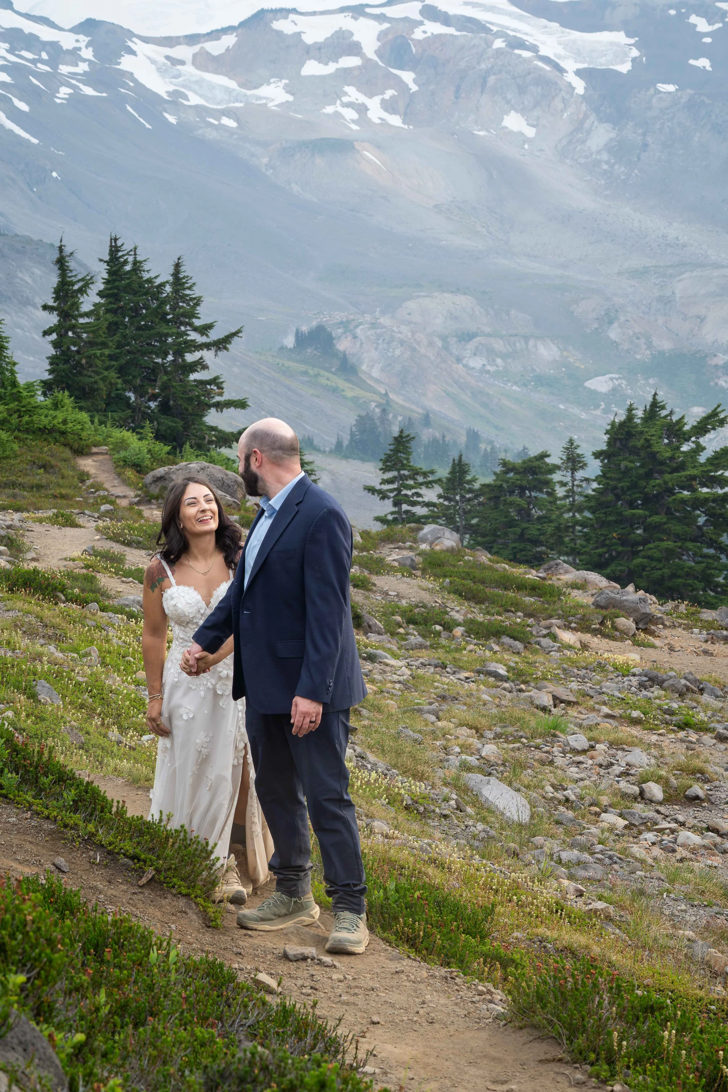 A bride and groom holding hands and walking through a subalpine meadow with blooming heather during their Ptarmigan Ridge elopement. The glaciated slopes of Mt. Baker are visible in the background.