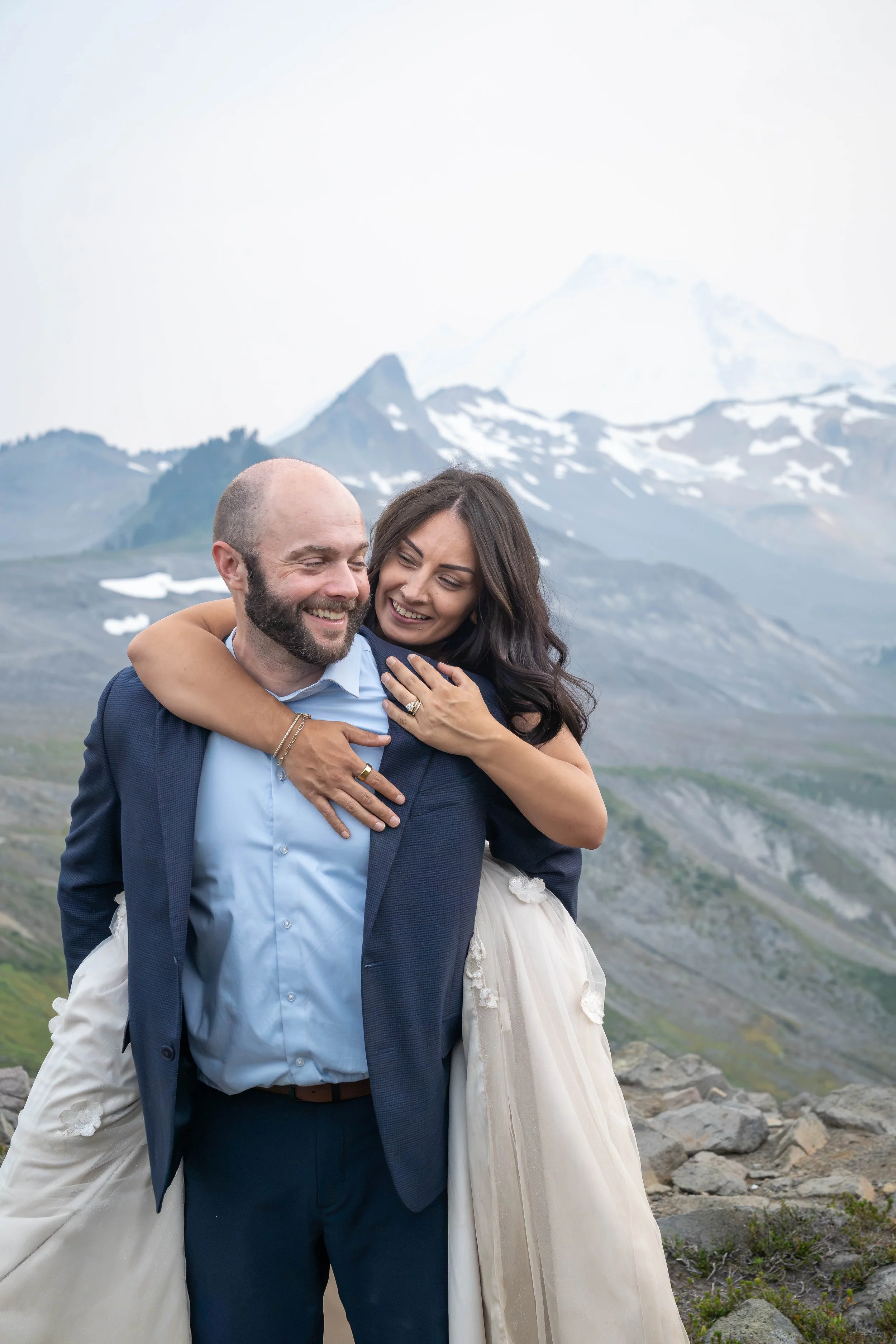 A bride jumps on her groom's back during their Ptarmigan Ridge elopement at sunset. It's hazy from wildfire smoke, giving the landscape a dreamy feel. Coleman Pinnacle and Mt. Baker are visible in the background.
