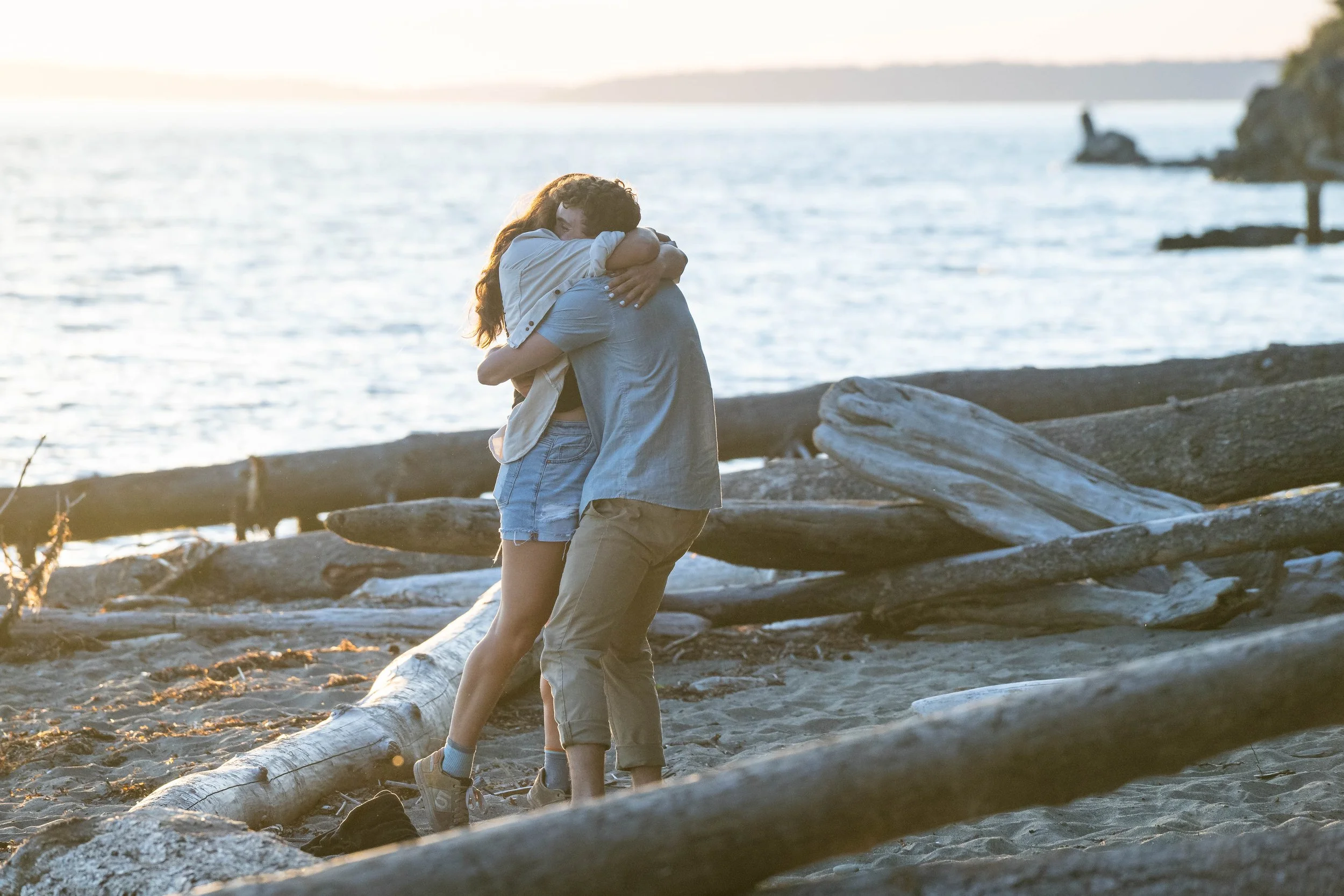A coupe hugging on Clayton Beach after getting engaged. They are in the sand surrounded by driftwood, with the ocean at sunset in the background.