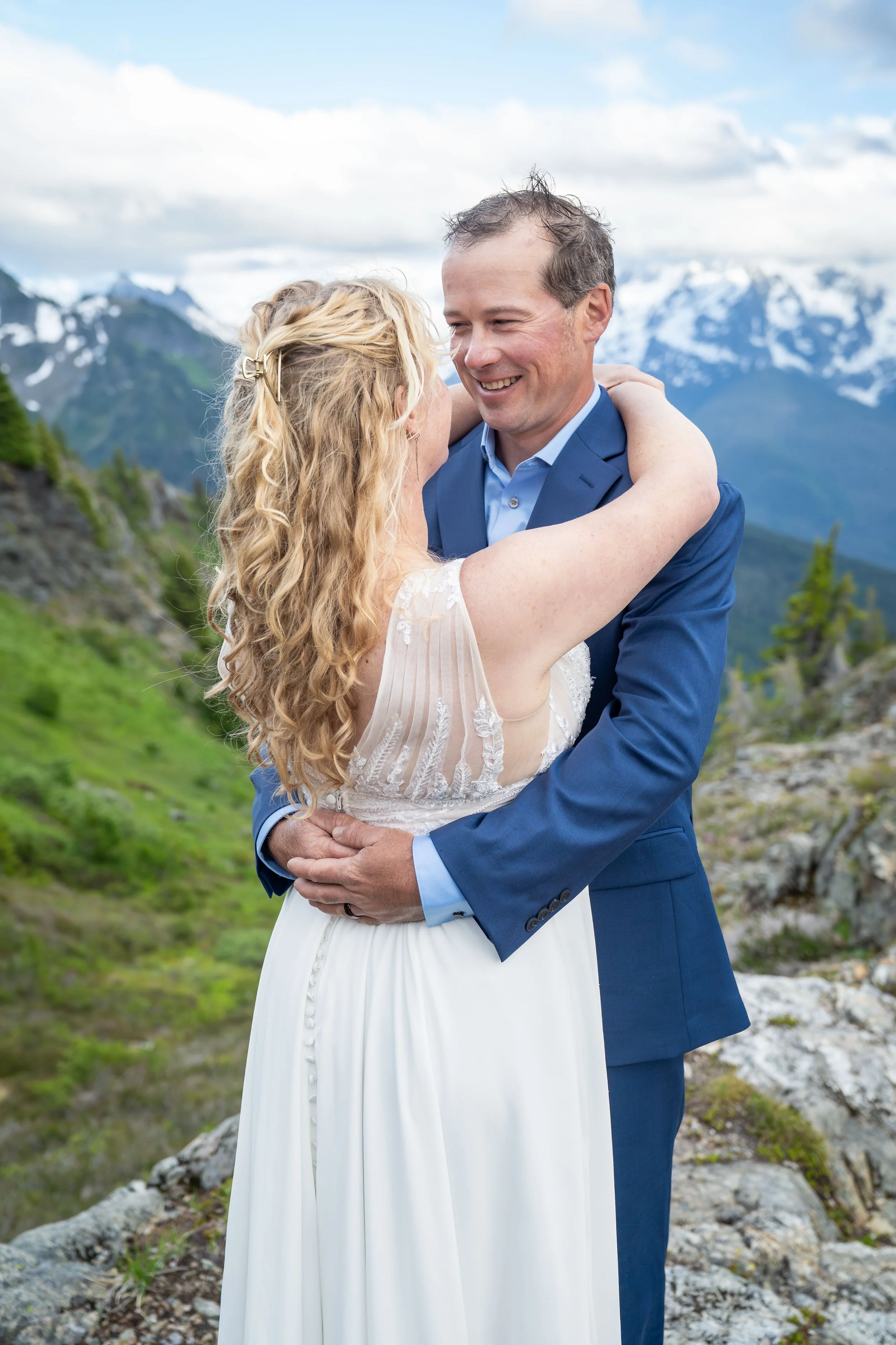 A bride and groom in wedding clothes holding each other and smiling during a North Cascades elopement at Yellow Aster Butte, with Mt. Shuksan visible behind them. They are standing on a rocky outcrop in a subalpine meadow.