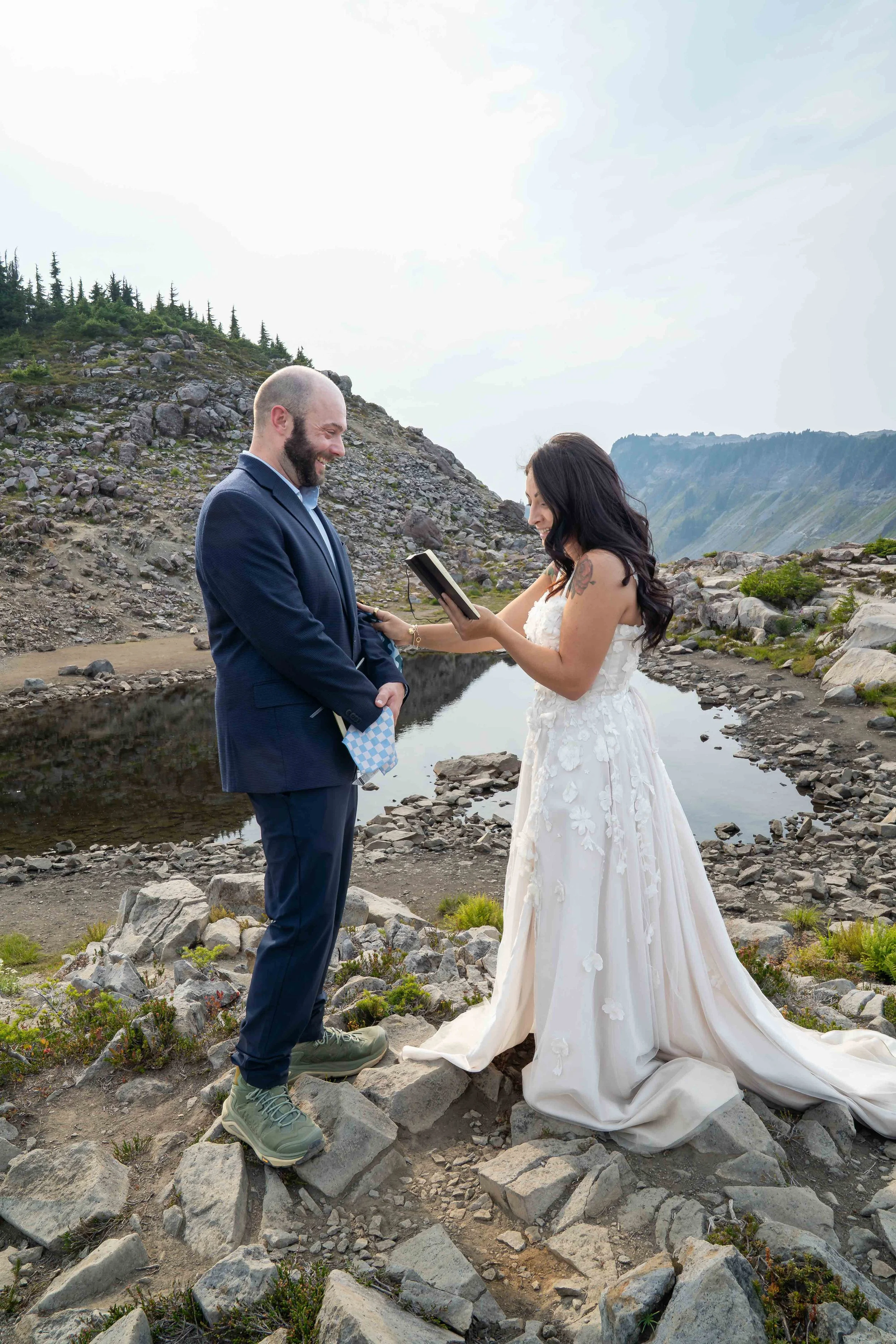 A bride reads her vows during an adventure wedding at Artist Point, near Mt. Baker. The bride and groom are standing in a rocky meadow with an alpine lake and Table Mountain behind them.