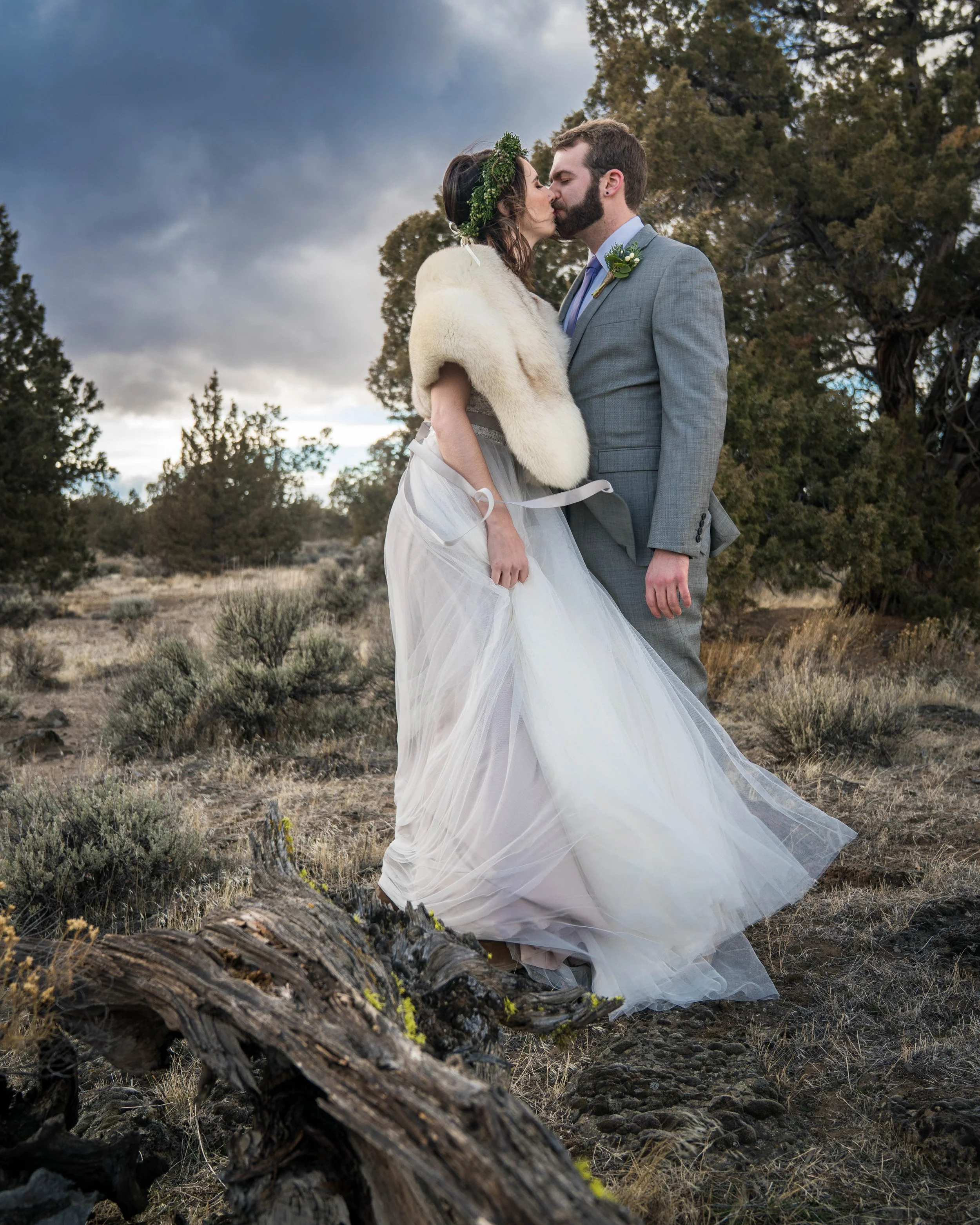 A photo of a bride and groom in wedding clothes standing together and kissing. They are in a desert landscape surrounded by sagebrush and the wind is blowing the bride's dress.