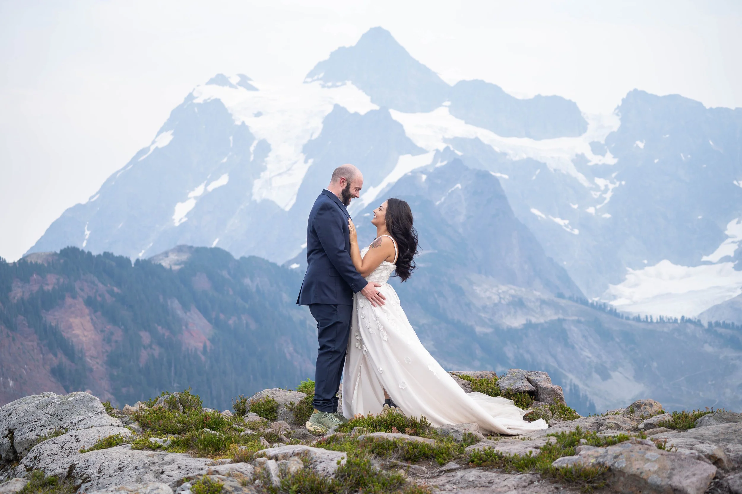 A bride and groom standing together beneath Mt. Shuksan and smiling at one another during their Artist Point elopement. The bride's white dress flows out from behind her.