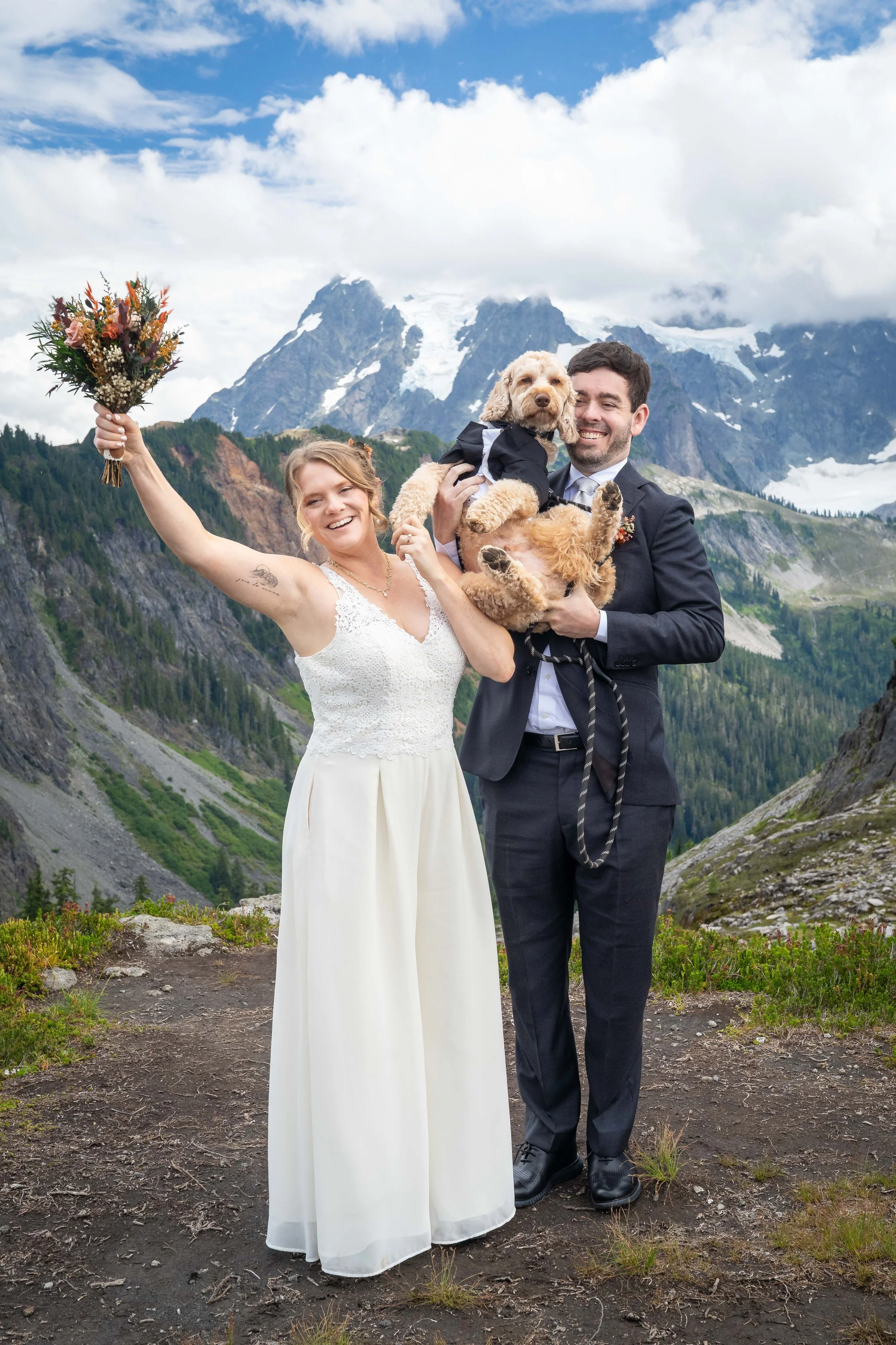Photo of a couple in a suit and wedding dress. The bride is holding a bouquet and the groom is holding a dog in a tuxedo.
