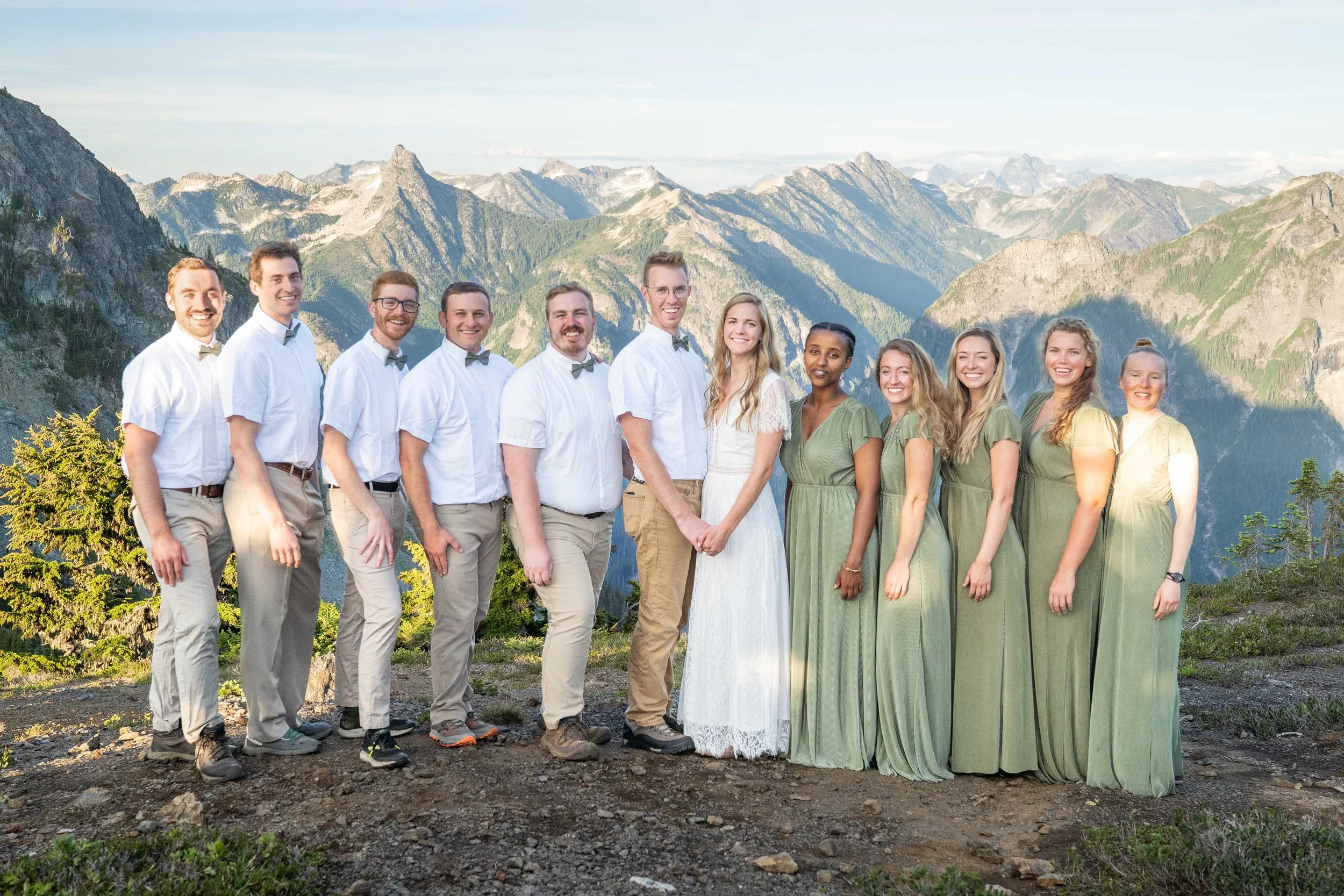 A group photo of a wedding party including a bride, groom, bridesmaids, and groomsmen during a North Cascades elopement. The bridesmaids and groomsmen are arranged around the couple and mountains are visible in the background.