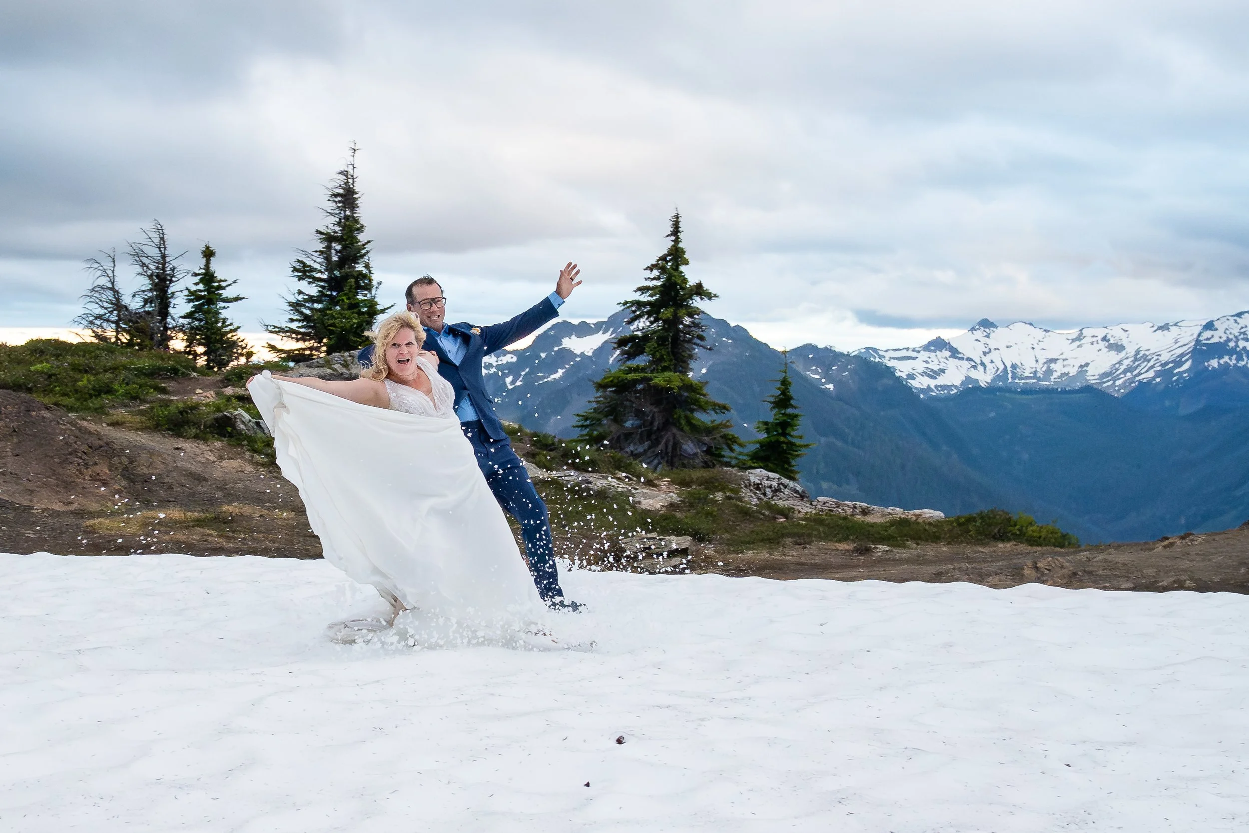 A bride in a wedding dress and a groom in a suit sliding on snow and holding their arms up during their North Cascades elopement at sunset. The snow-covered slopes of Mt. Baker are visible in the background.