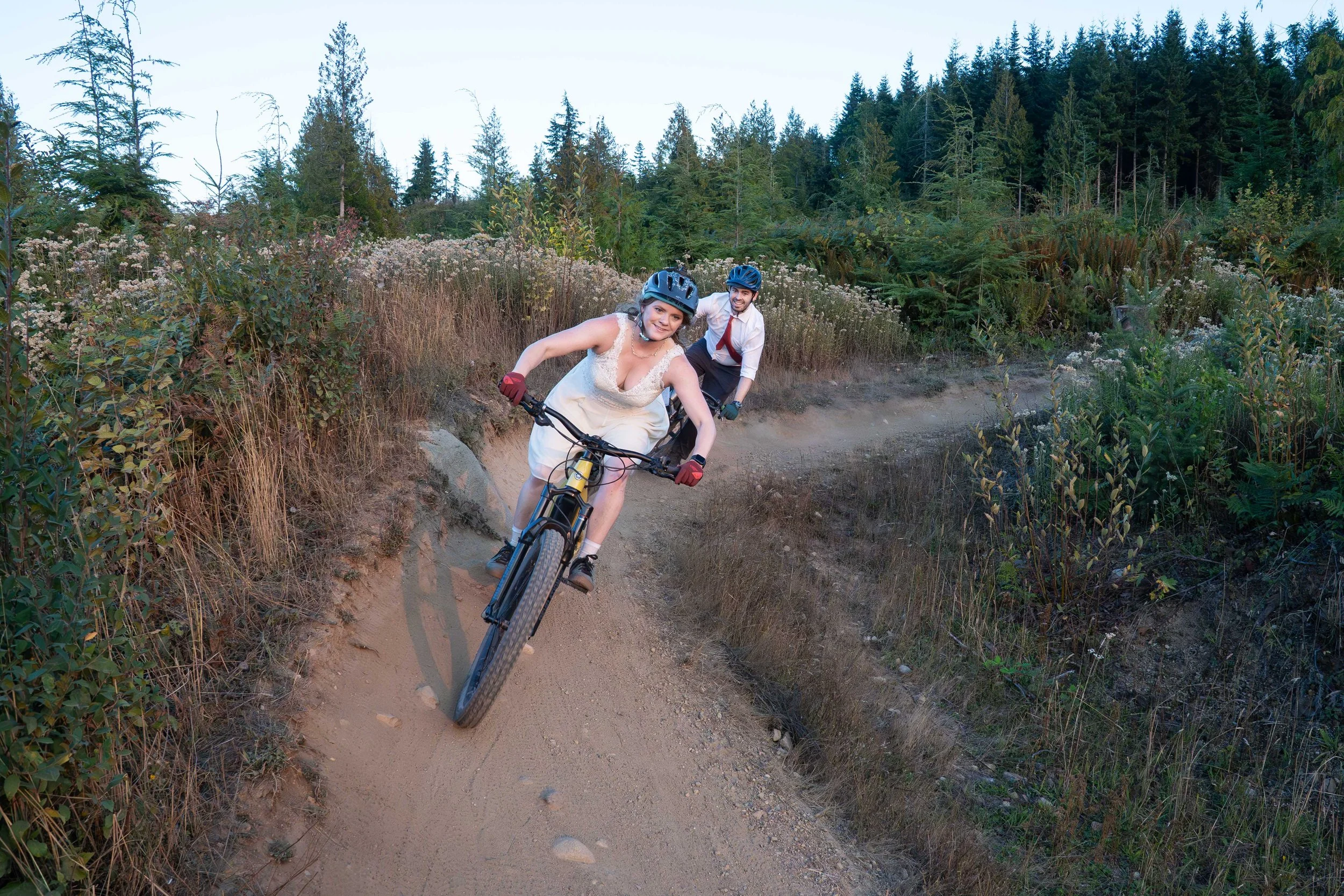 A couple in wedding clothes riding mountain bikes down SST on Galbraith Mountain at sunset during their mountain biking elopement.