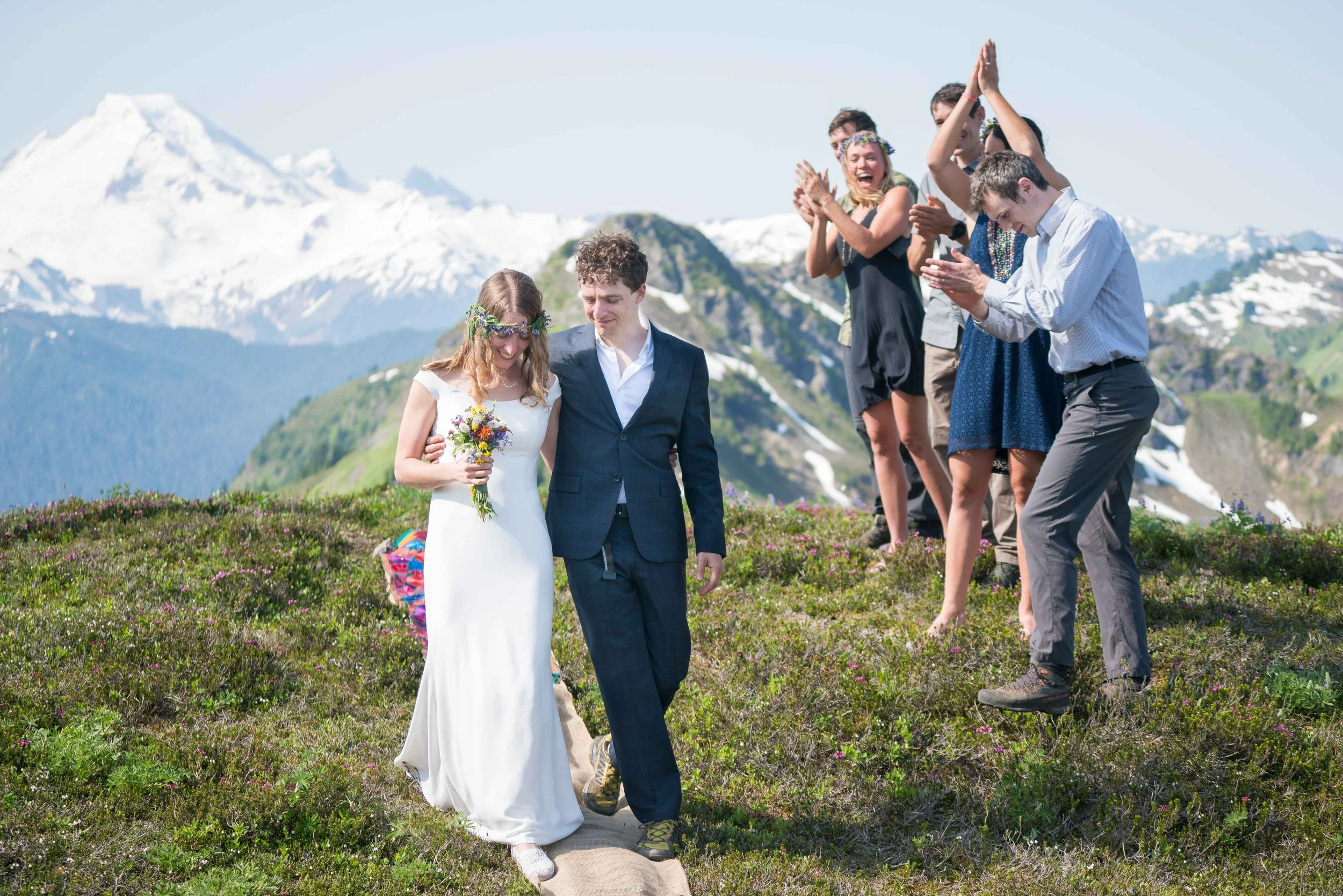 A couple in wedding clothes walks down a makeshift "aisle" as their friends clap and cheer after a backpacking elopement in the North Cascades. Mount Baker is visible in the background.